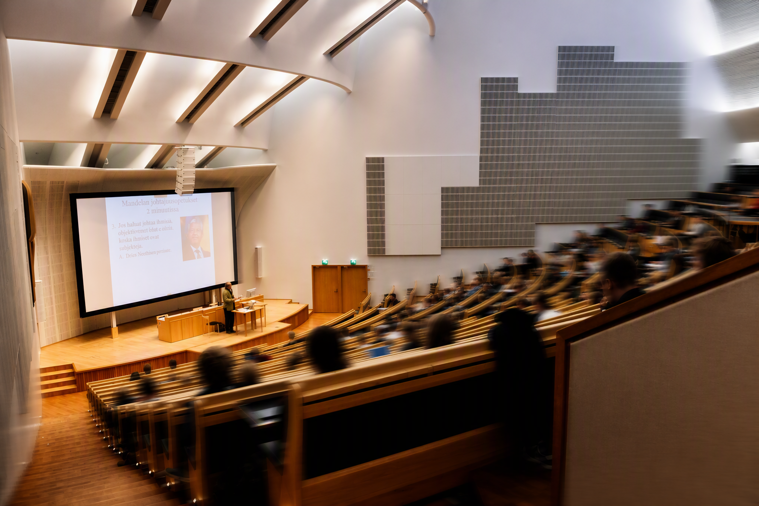 A large lecture hall with a presenter standing at the front near a podium and a wide screen displaying a presentation slide featuring text and a photo of a man, with attendees seated in tiered wooden benches.