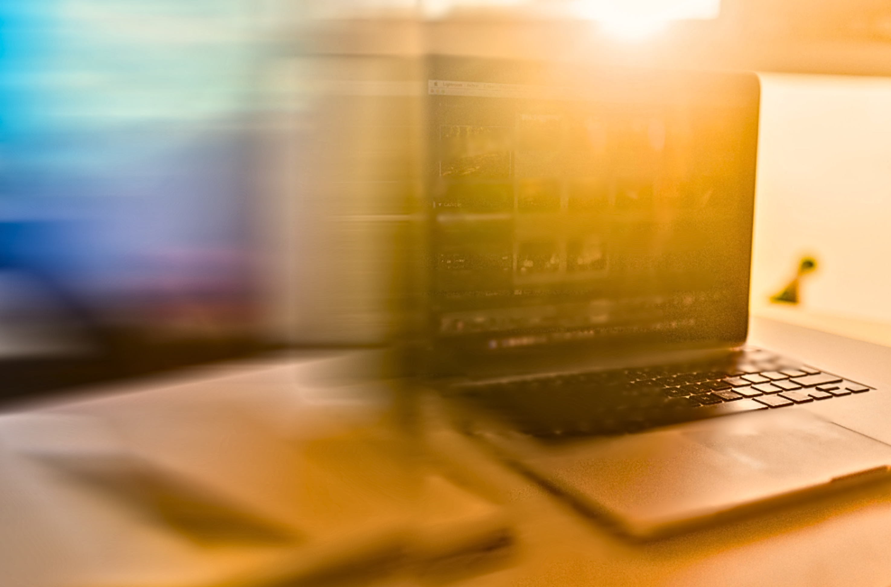 Partially blurred image of a laptop on a desk with soft warm and cool lighting, showing a faint screen displaying an open photo editing software.