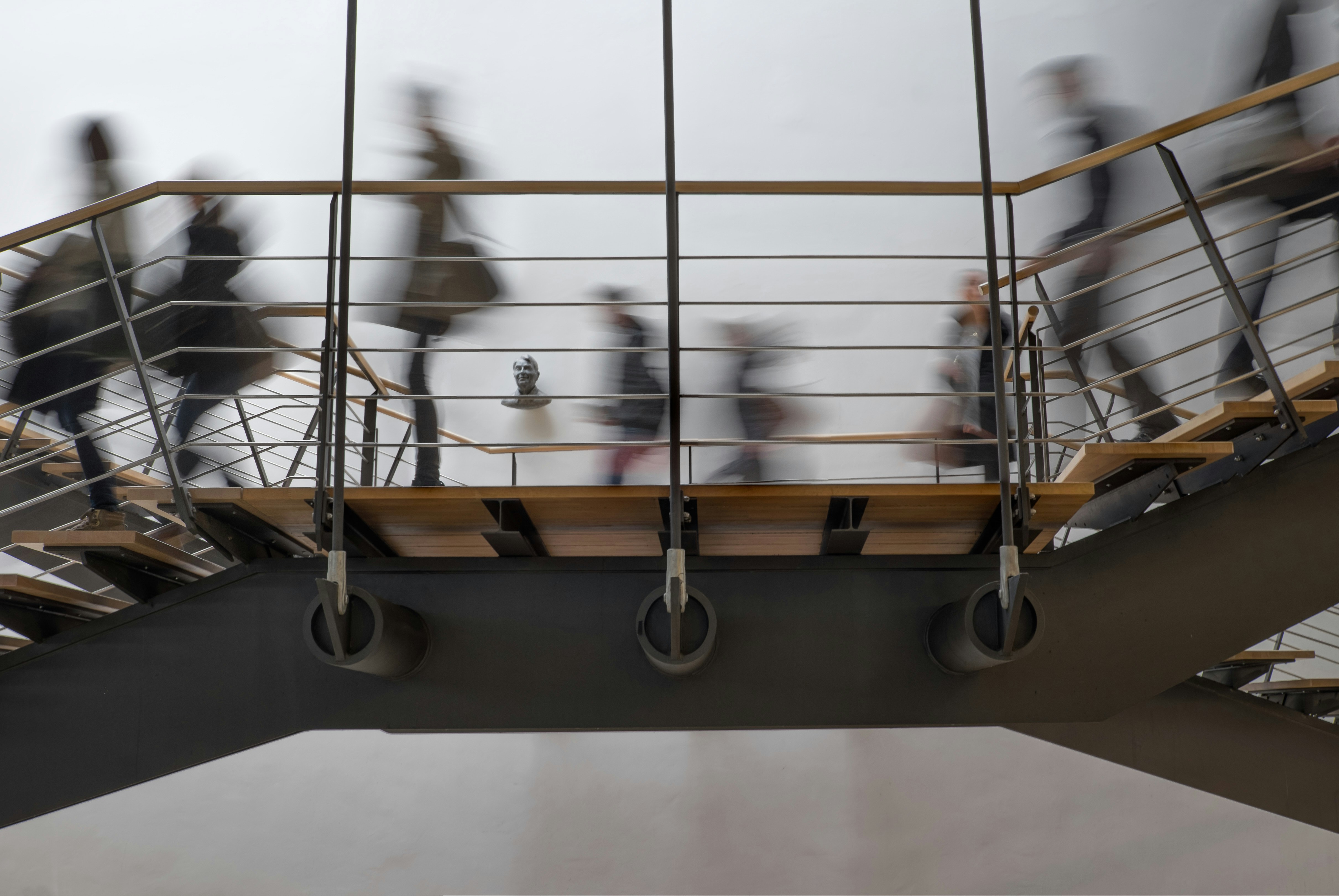 Blurred figures of people walking on a modern metal and wood staircase against a plain wall with a small bust sculpture mounted on it.