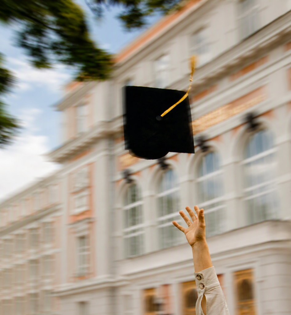Hand raised tossing a black graduation cap with a yellow tassel in the air outside a classical building with large windows.