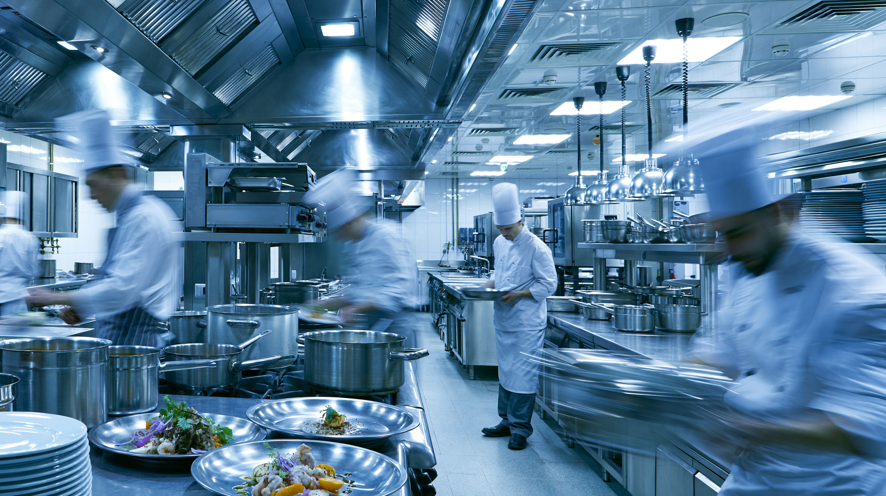Busy professional kitchen with chefs in white uniforms and tall hats preparing dishes amid stainless steel pots, pans, and plates, showing motion blur of activity.