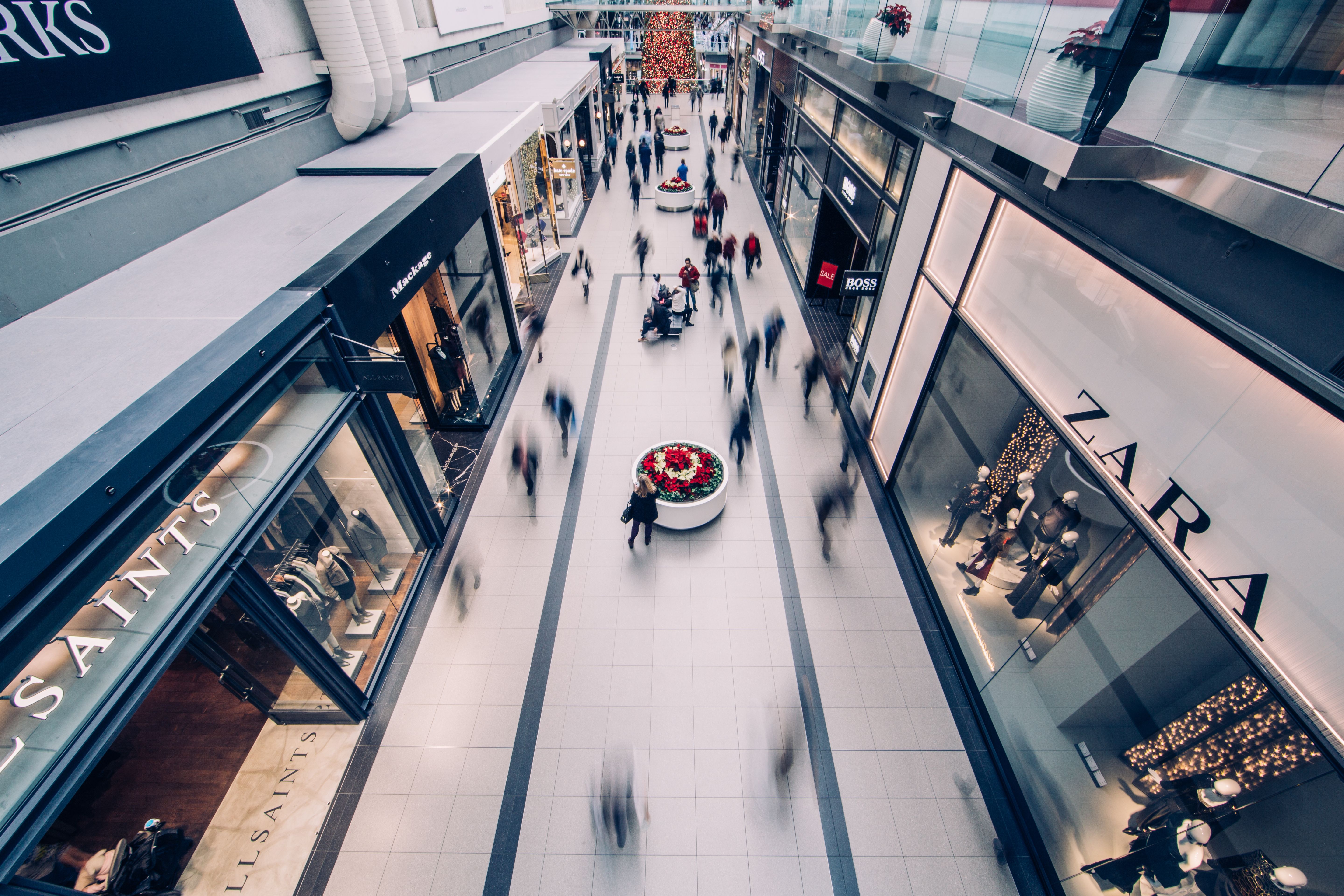 Overhead view of a shopping mall walkway with blurred shoppers moving past stores including Zara and AllSaints, with festive red flowers in planters along the center.