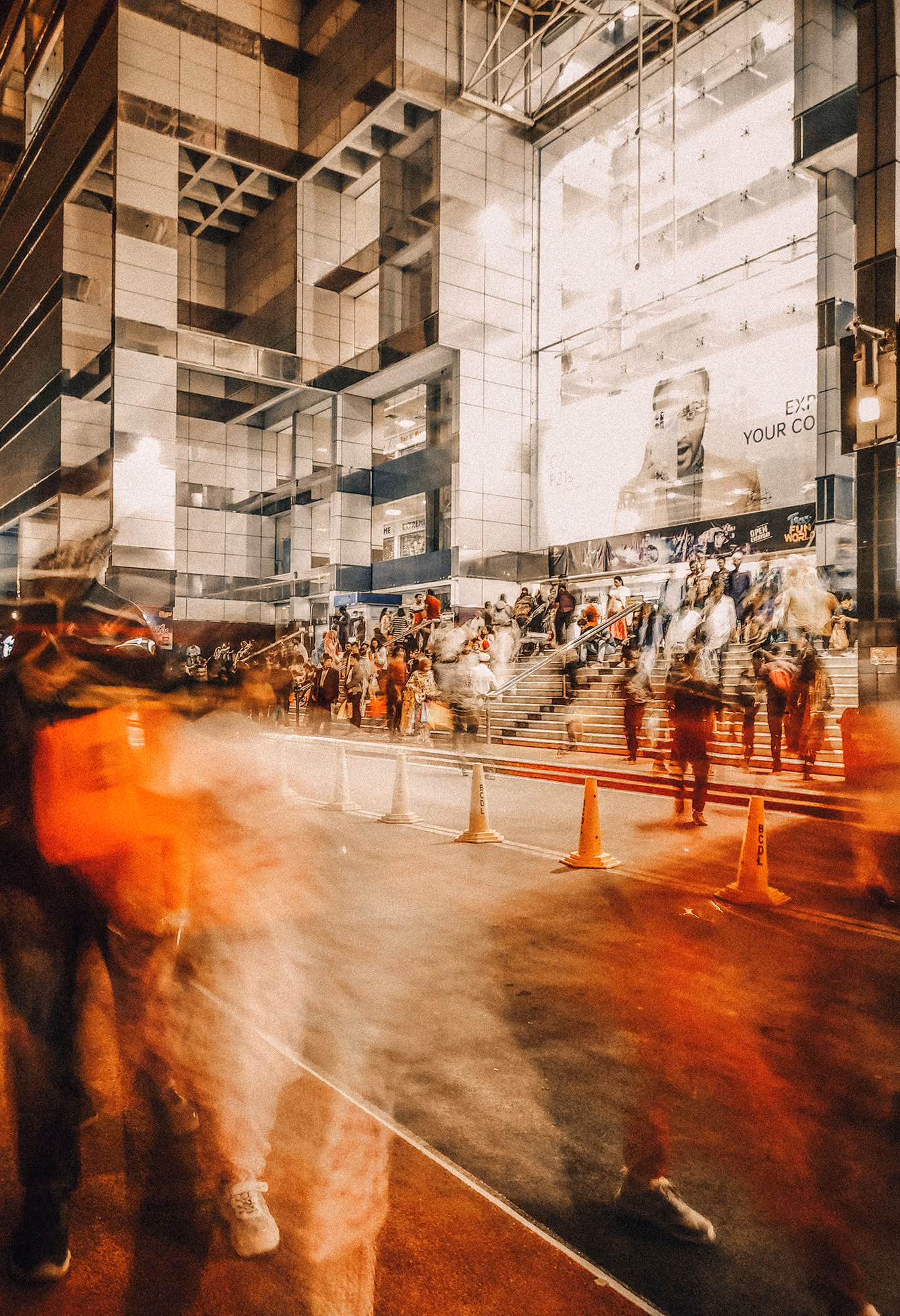 Blurred crowd of people walking and gathered near steps outside a modern building with reflective glass and a large billboard.