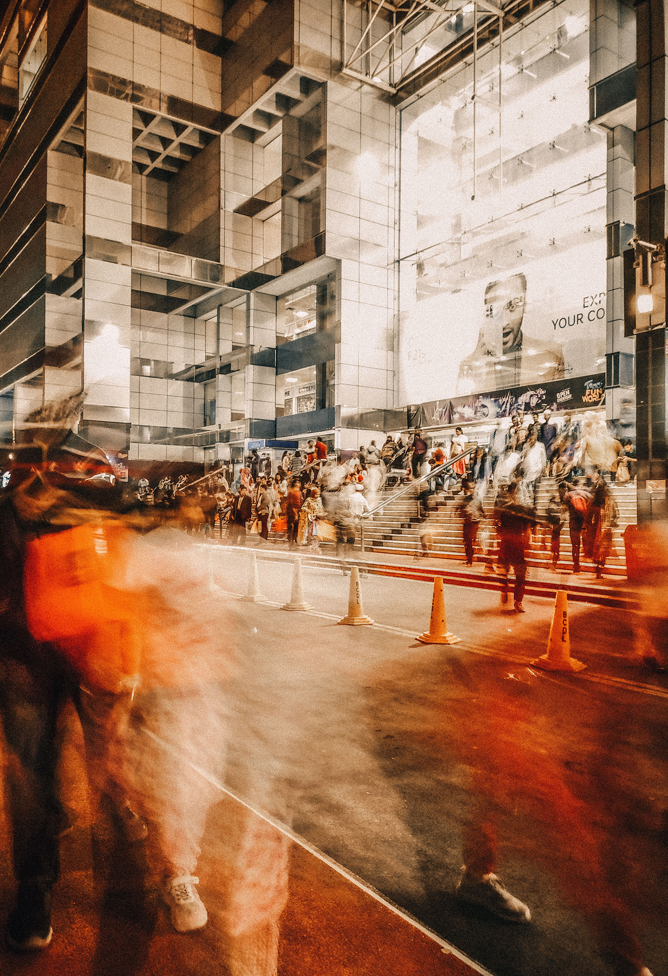Blurred crowd of people walking and gathered near steps outside a modern building with reflective glass and a large billboard.