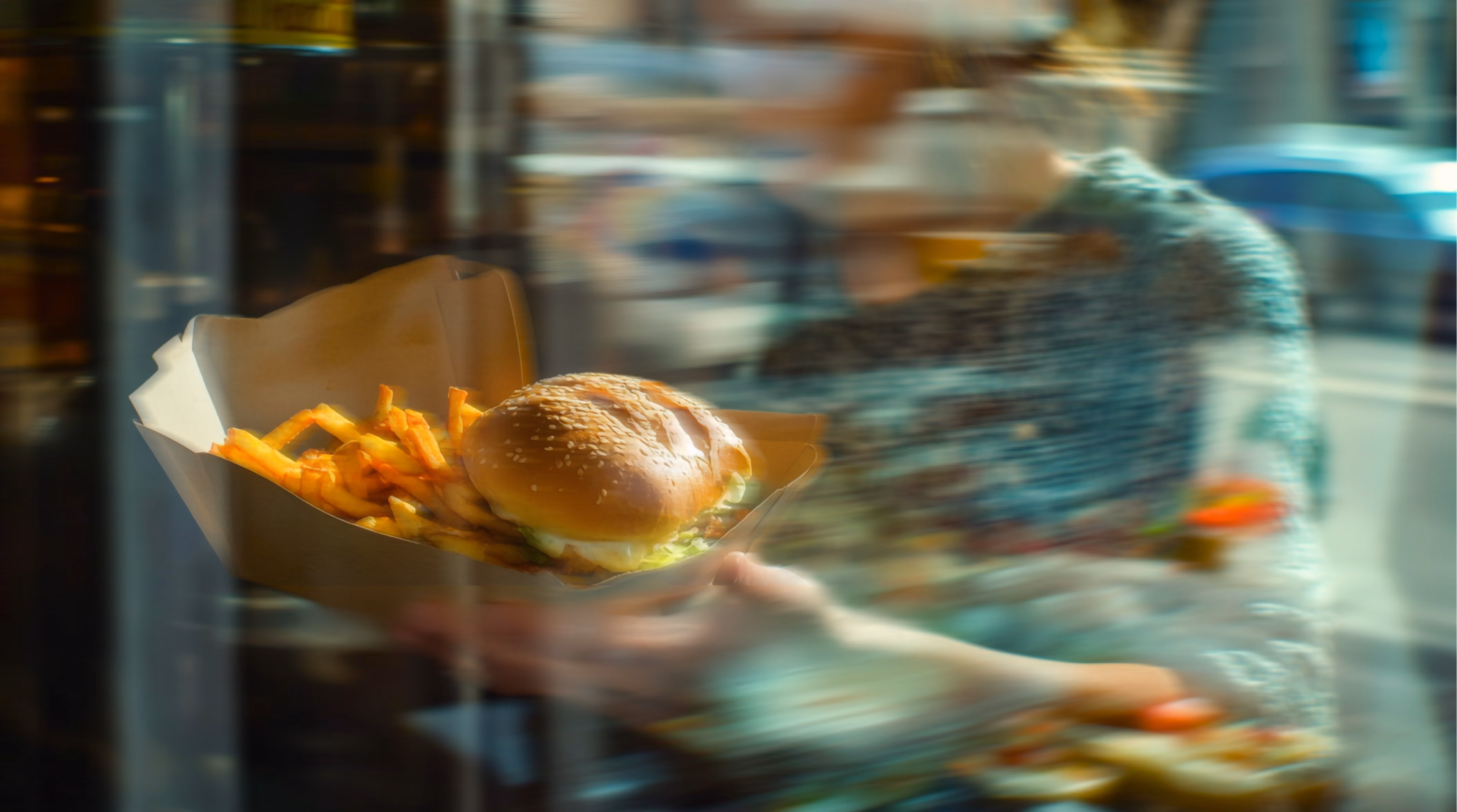 Person holding a cardboard tray with a cheeseburger topped with lettuce and a serving of French fries, seen through a window with reflections and blur effects.
