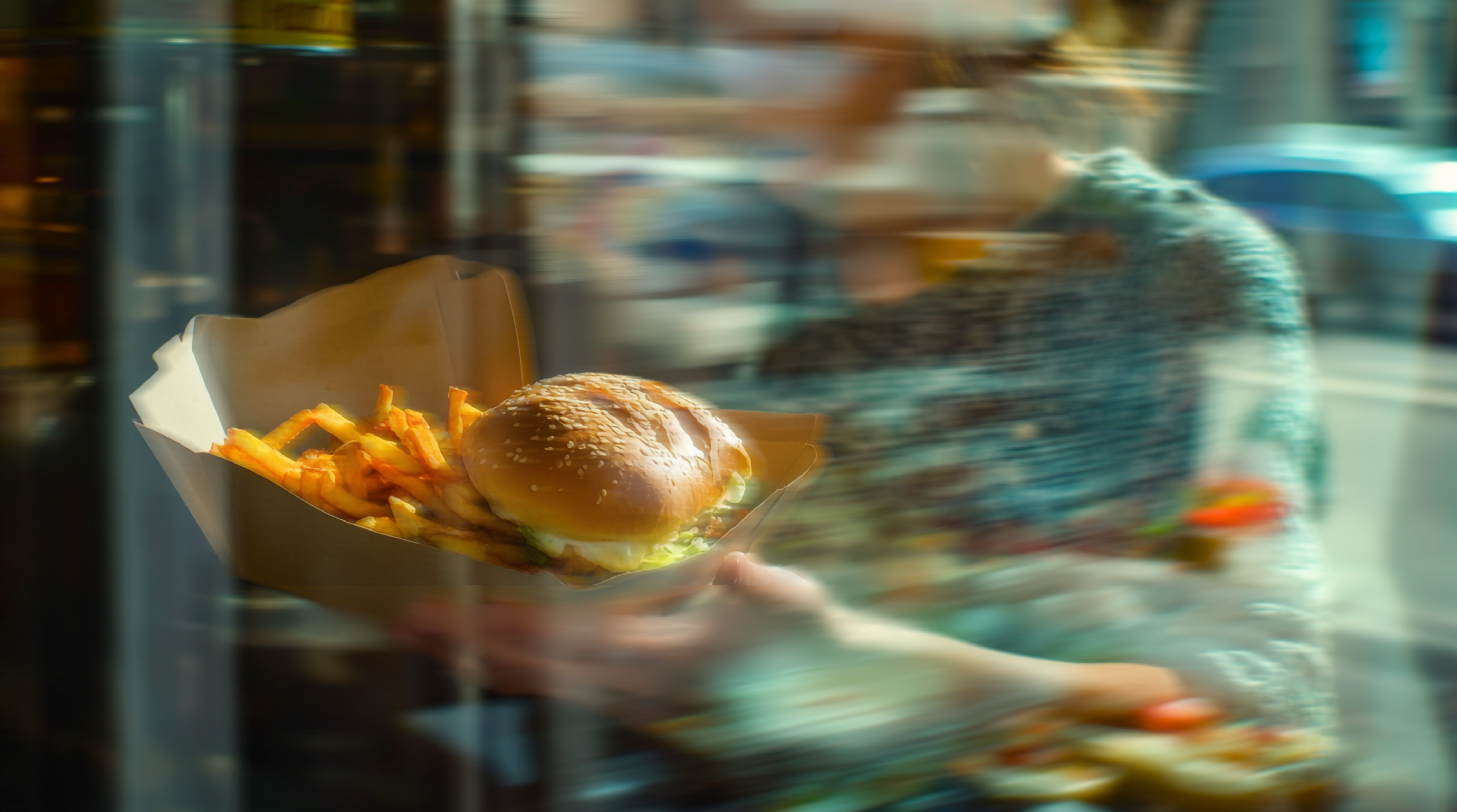 Person holding a cardboard tray with a cheeseburger topped with lettuce and a serving of French fries, seen through a window with reflections and blur effects.