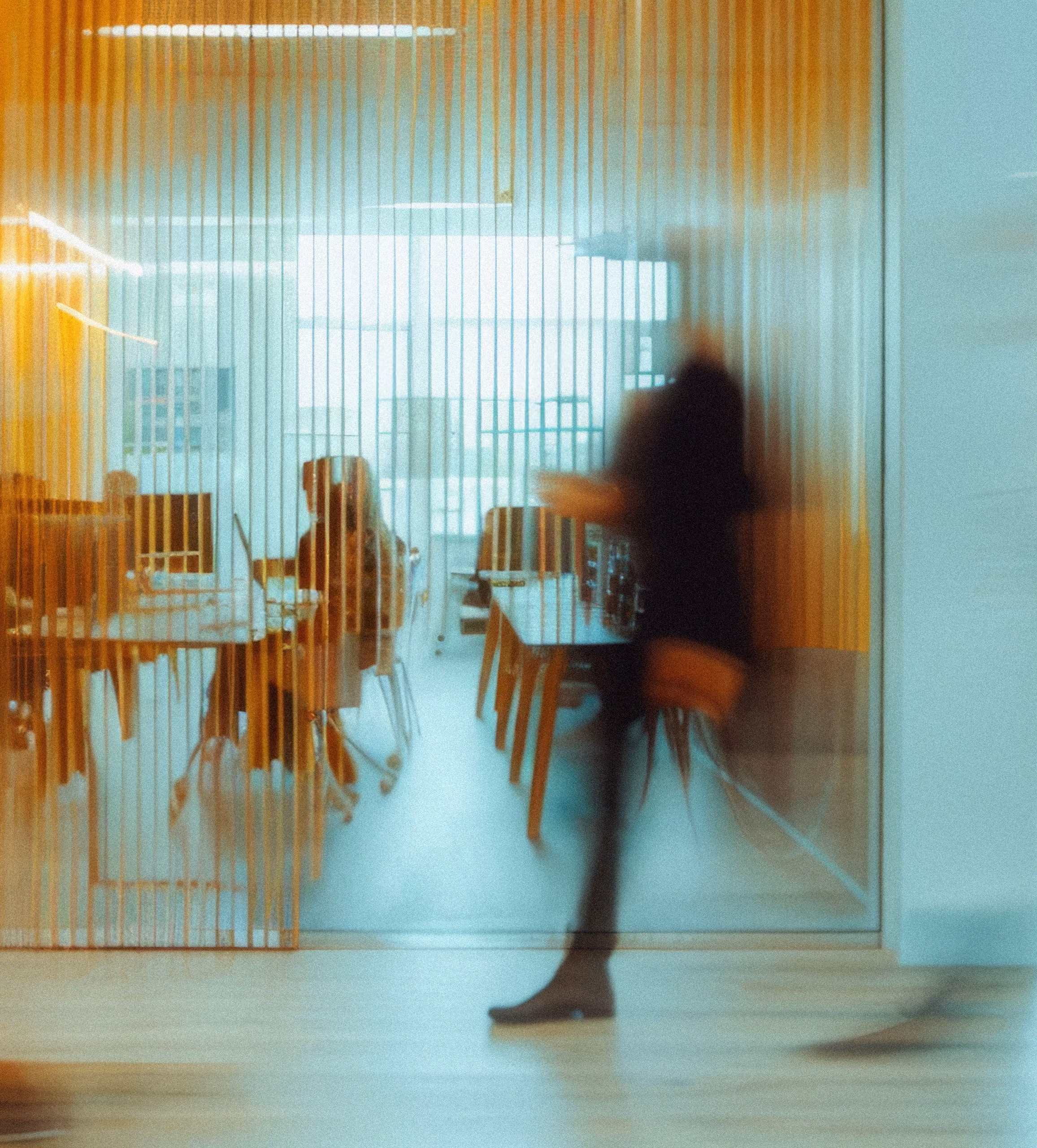 Blurred image of a person walking past a glass office with vertical yellow blinds; inside, two people sit at desks with computers in a modern office space.