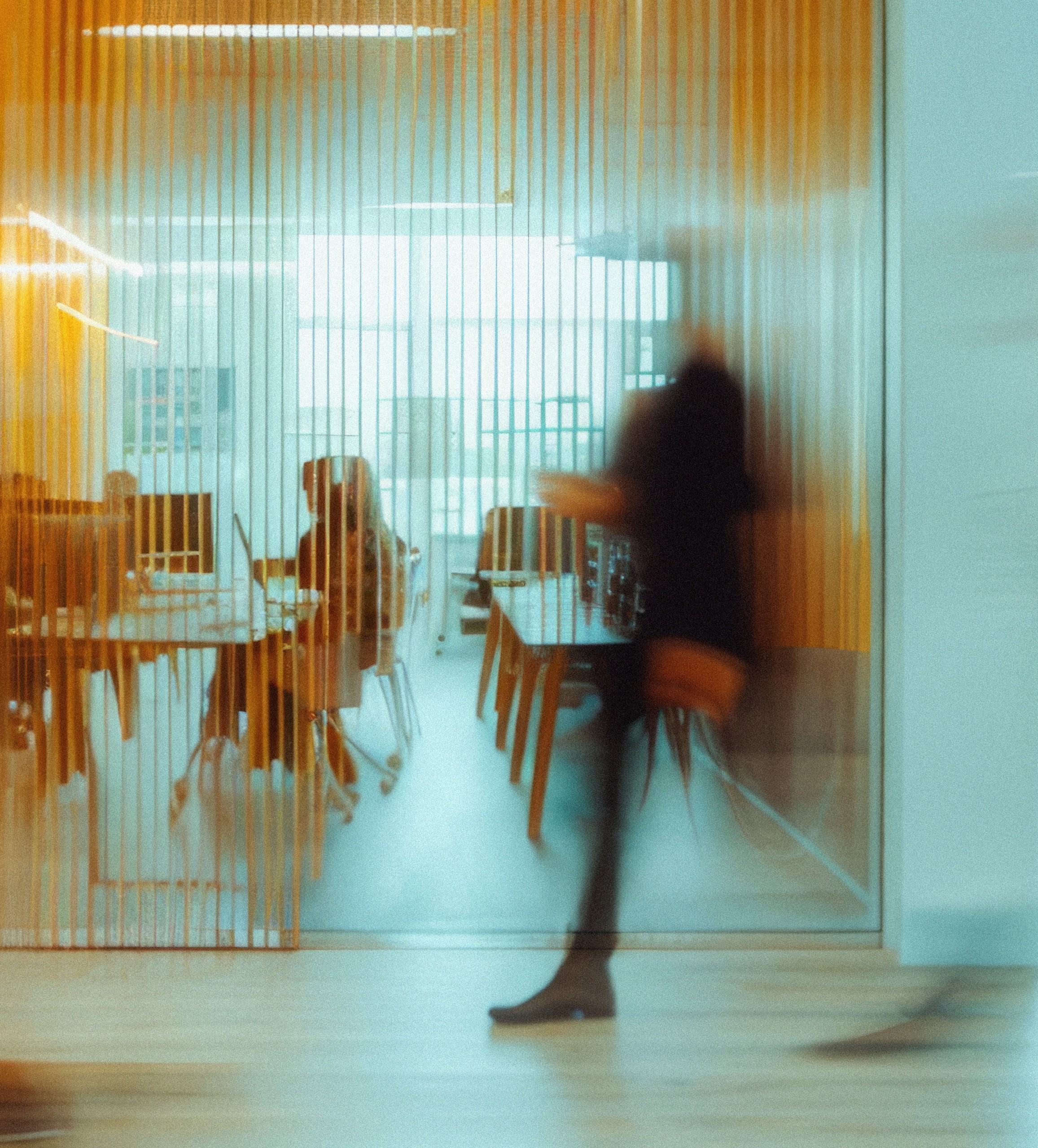 Blurred image of a person walking past a glass office with vertical yellow blinds; inside, two people sit at desks with computers in a modern office space.