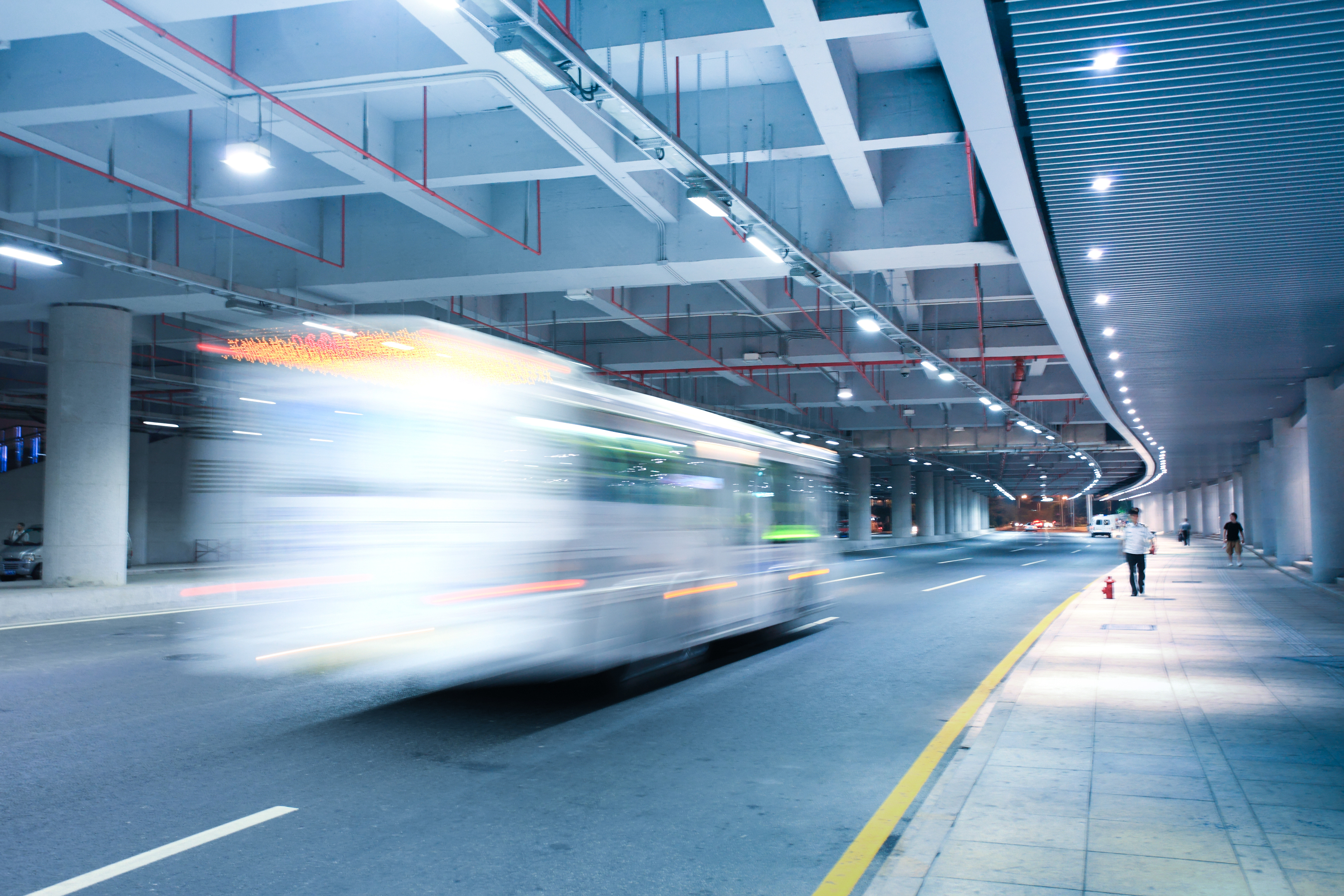 Blurred white bus moving quickly on a wide road under an illuminated modern overpass at night with a few pedestrians walking on the sidewalk.
