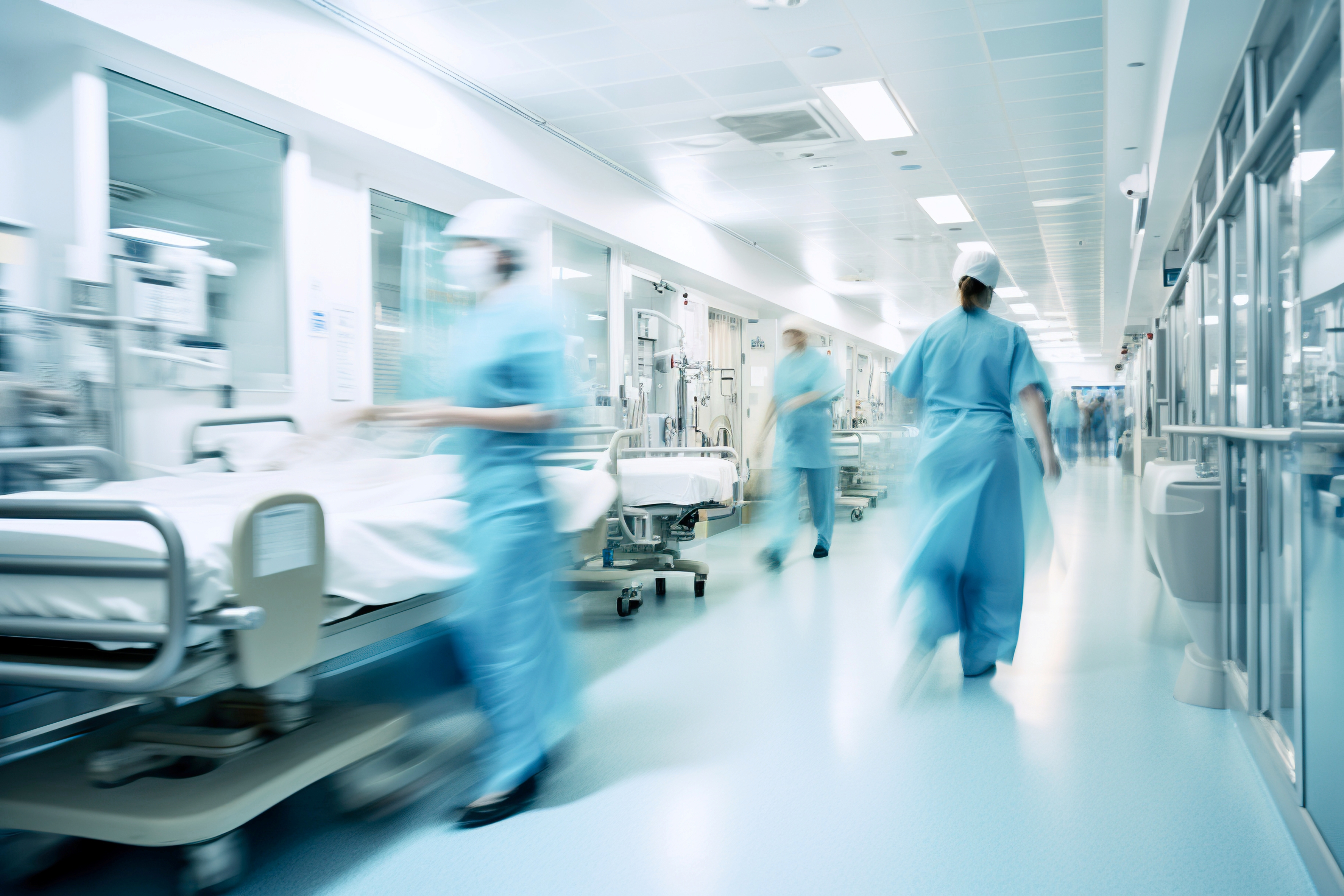 Blurred motion image of healthcare workers in blue scrubs and masks walking in a bright hospital hallway with empty patient beds lined against the walls.