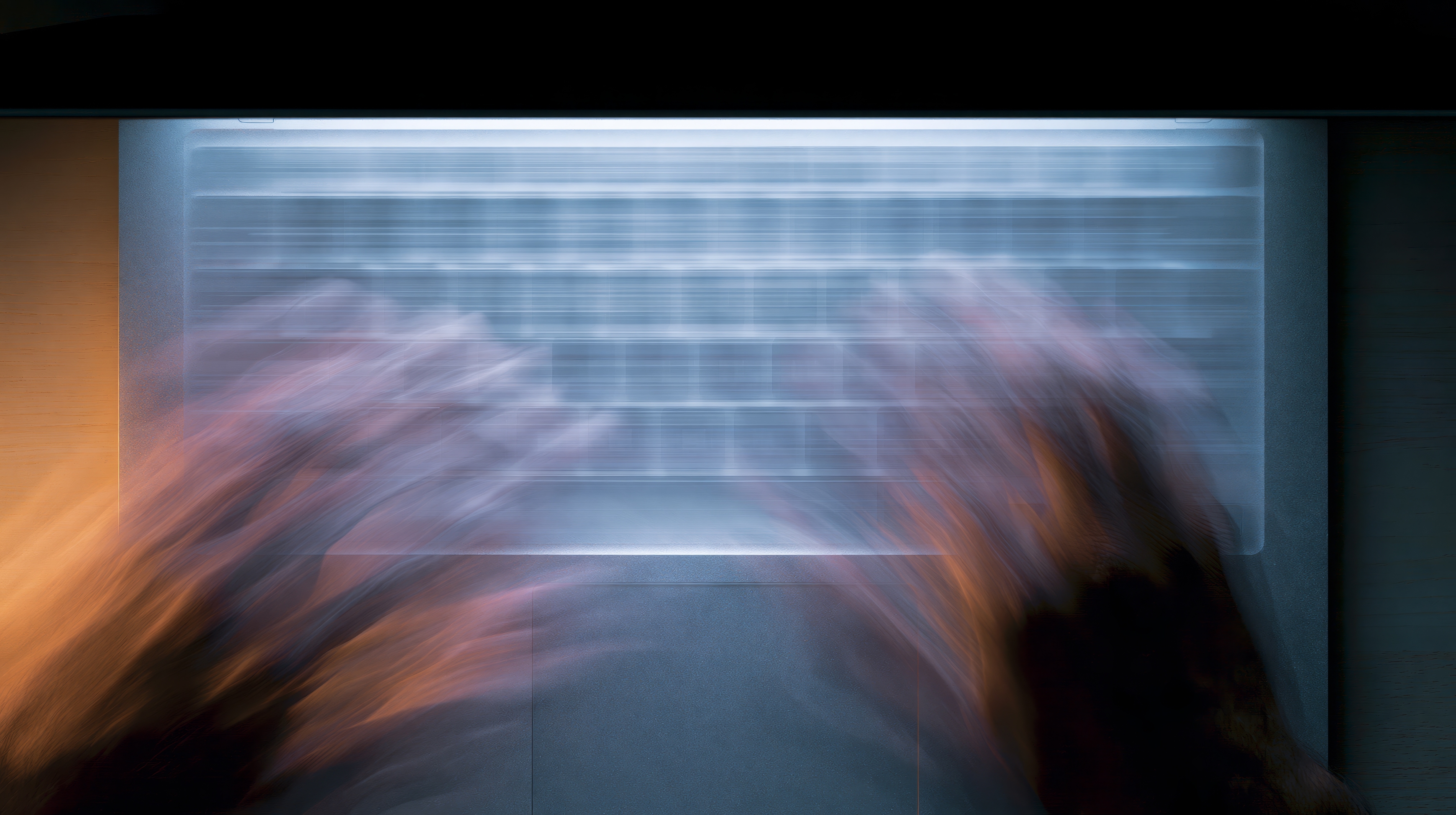 Blurred motion of hands typing rapidly on a laptop keyboard with a wooden desk beneath.