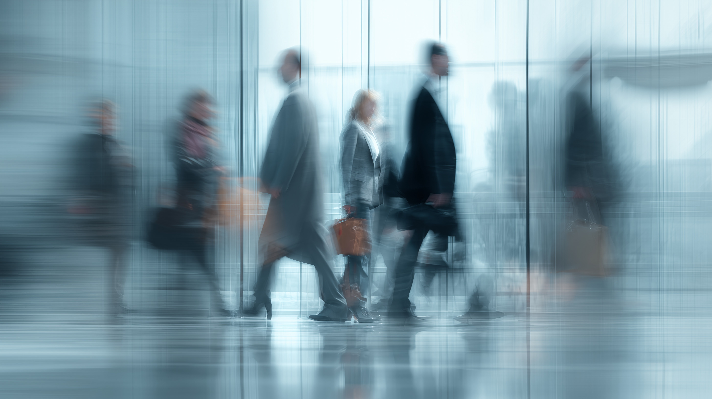 Blurred motion image of multiple business people walking indoors, carrying briefcases and bags, with reflections on the shiny floor and a blue tint.