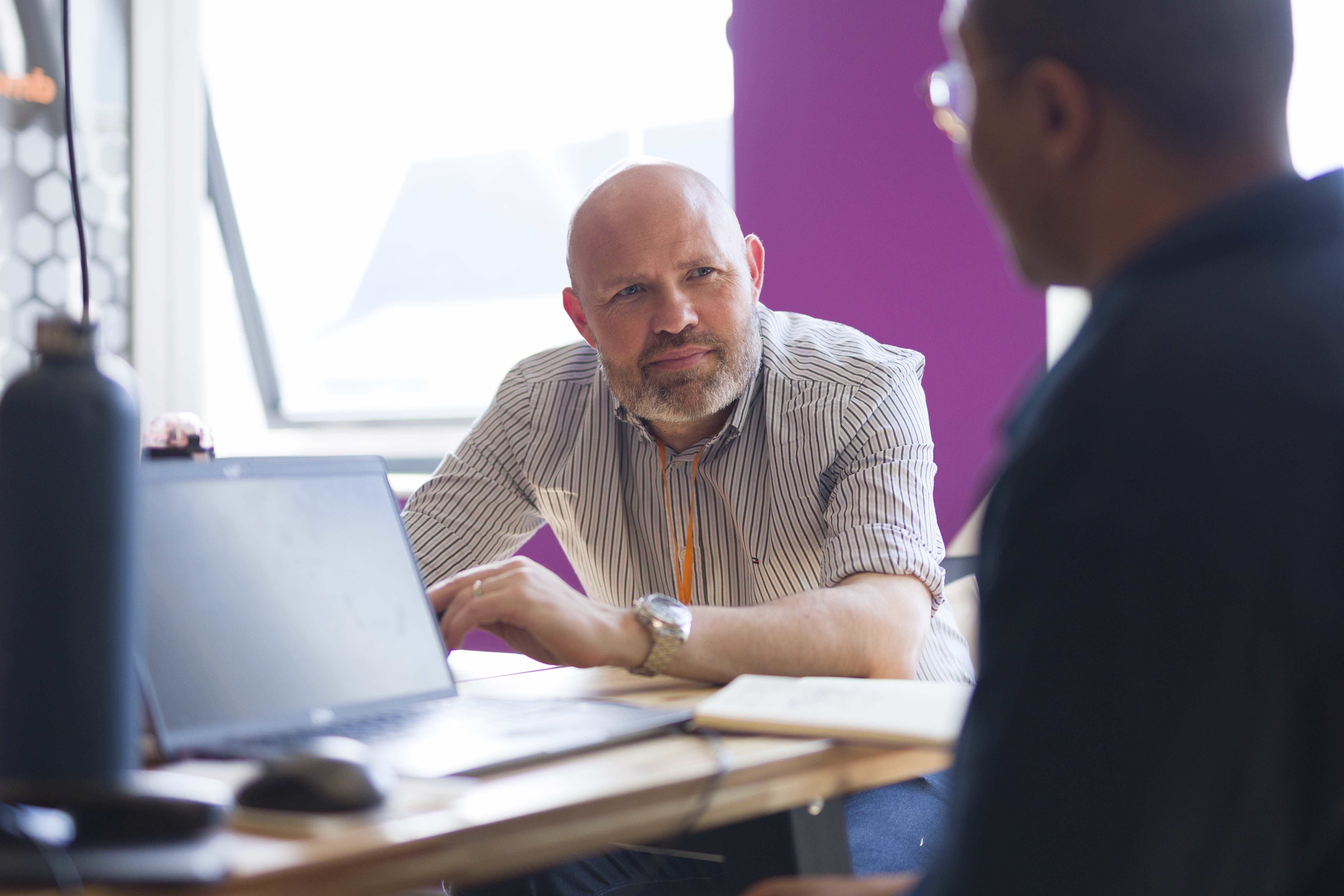 Two men engaged in a conversation at a desk in a bright office; one man with a bald head and beard wearing a striped shirt leans on the desk, facing the other man whose back is to the camera, while a laptop and notebook sit on the desk.