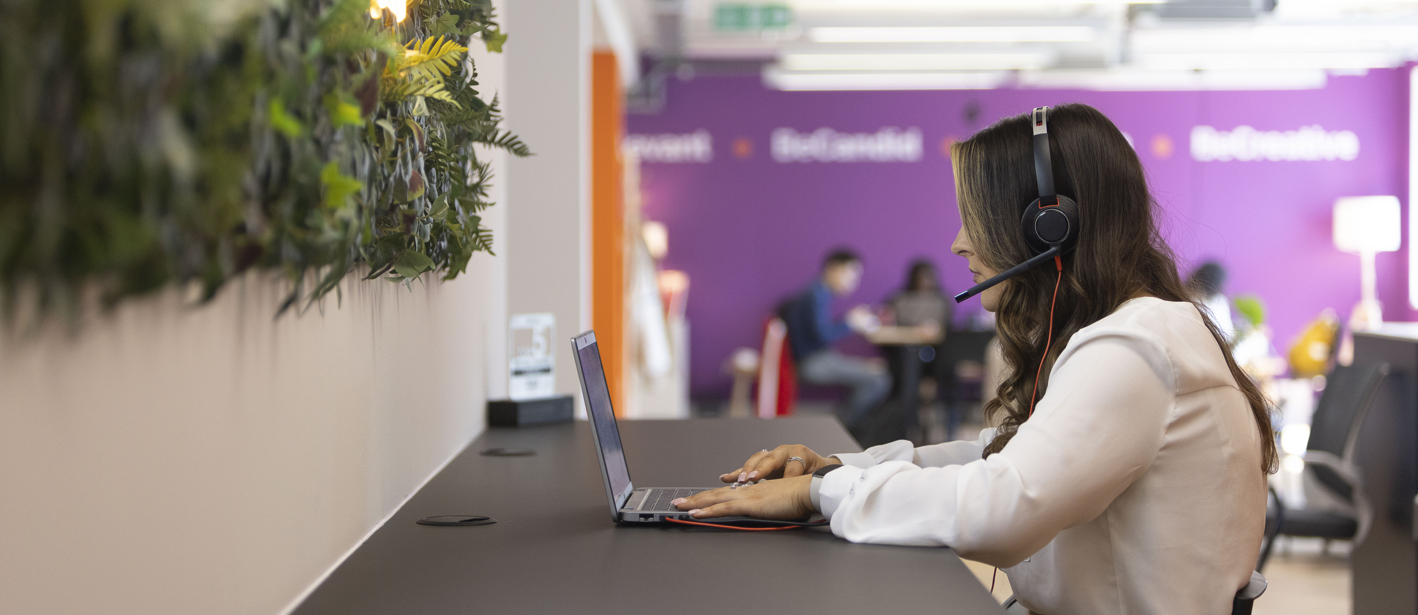 Woman with long brown hair wearing a white blouse and black headset working on a laptop at a counter in a modern office with a green plant wall on the left and a purple wall in the background.
