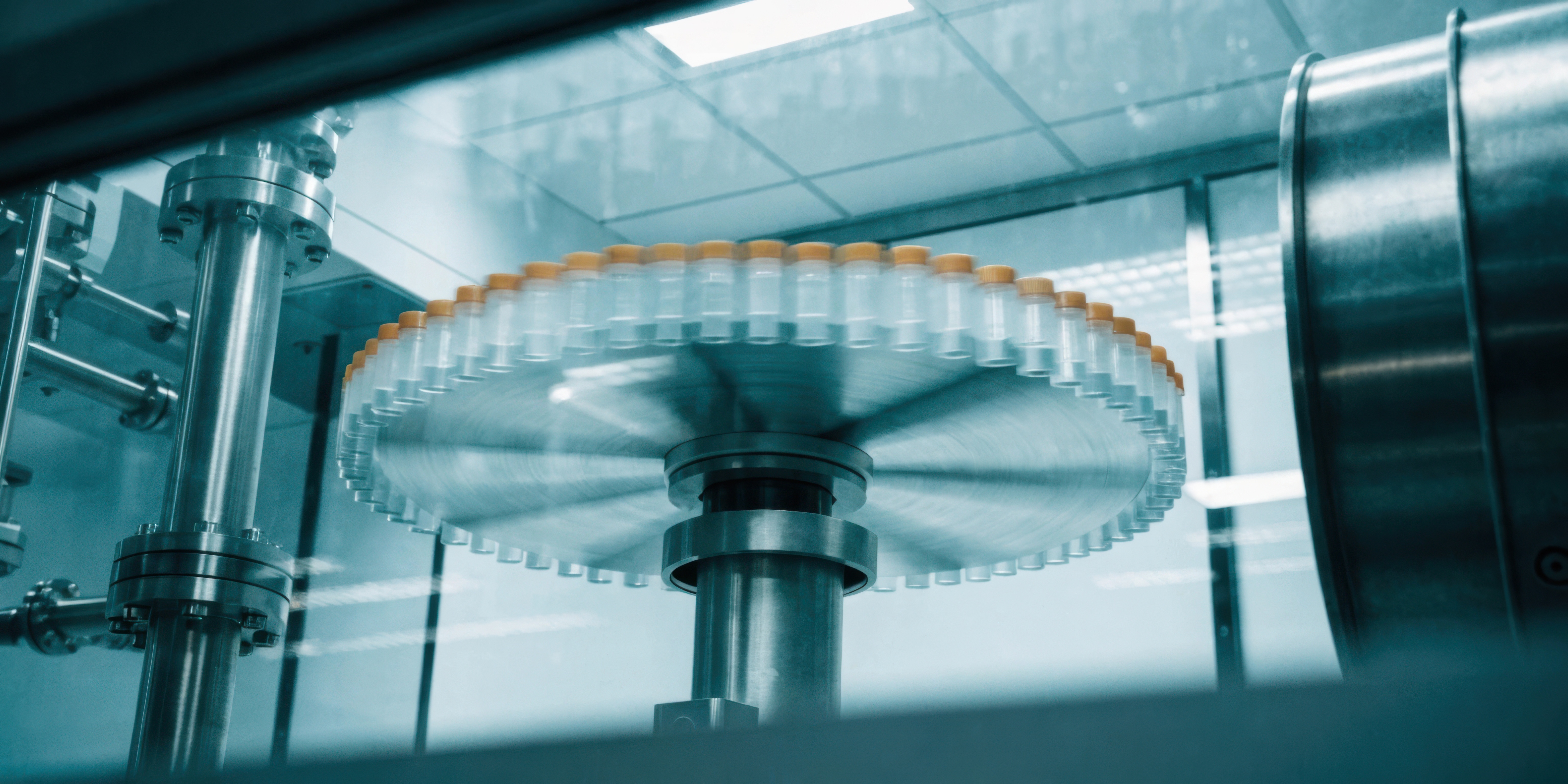 Close-up of a pharmaceutical production machine with a rotating circular platform holding multiple sealed glass vials with orange caps inside a clean, well-lit environment.