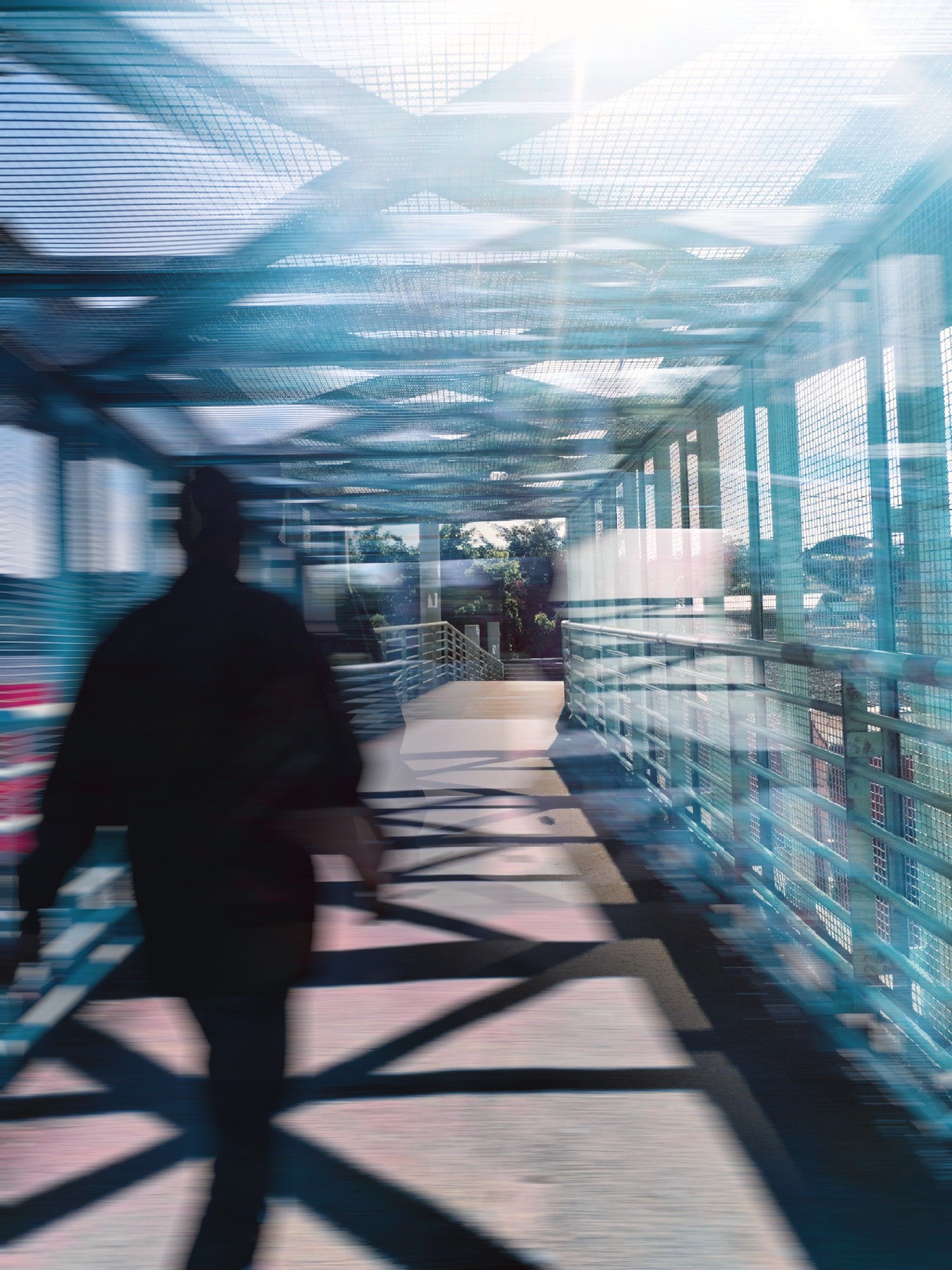 Silhouetted figure walking through a sunlit, mesh-covered bridge or walkway with blue metal framework casting shadows on the ground.