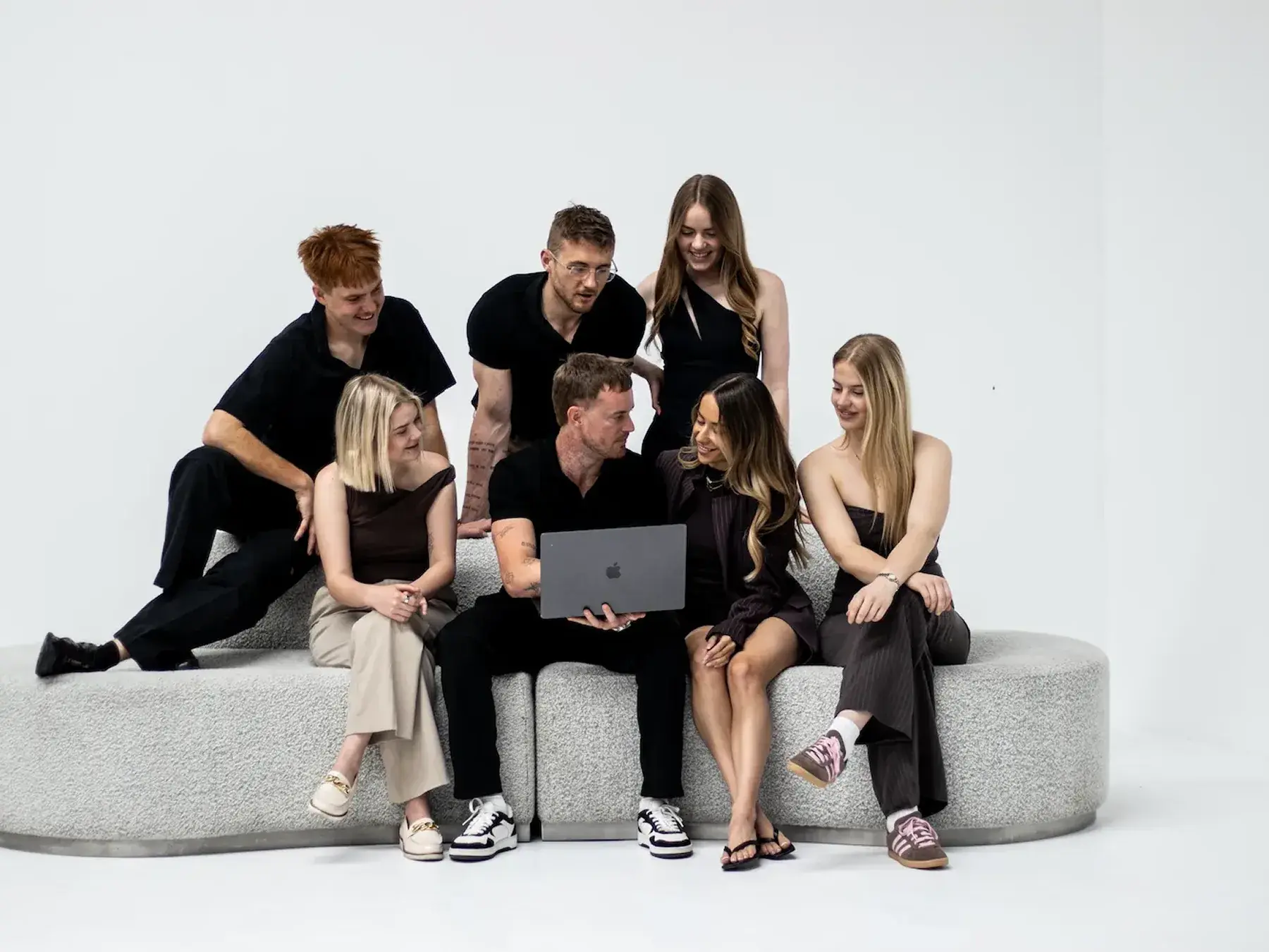 Seven young adults sitting and standing on a rounded gray couch looking at a laptop together.