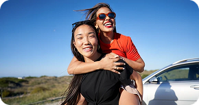 Two women outdoors smiling, one piggybacking the other near a white car under clear blue sky.
