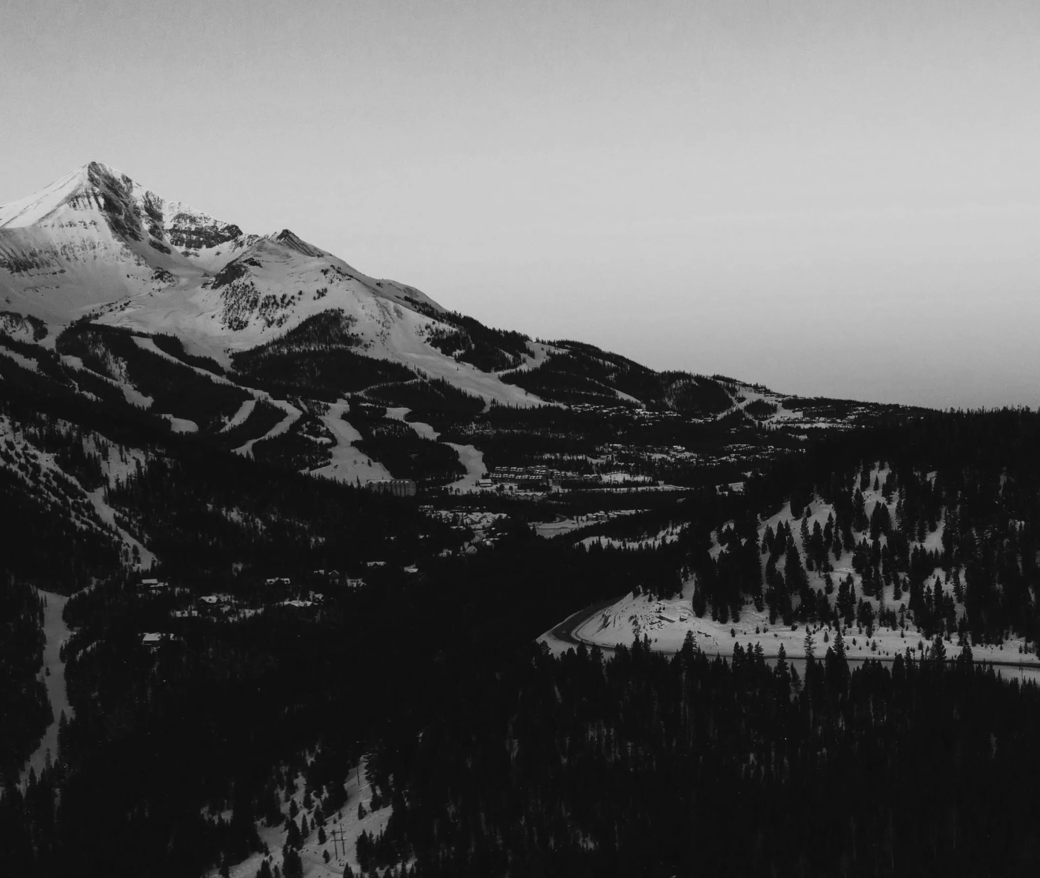 Snow-covered mountain and forest landscape with ski slopes and a winding road below.