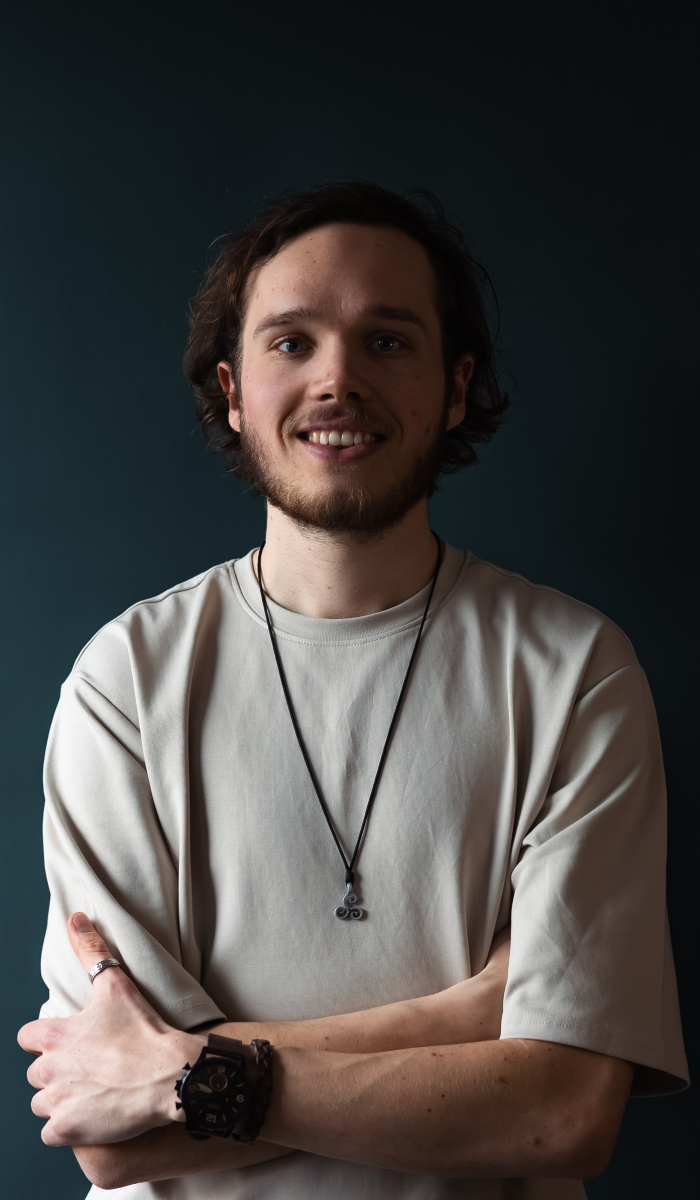 Smiling young man with beard wearing beige shirt, necklace, ring, and watch crossing arms.