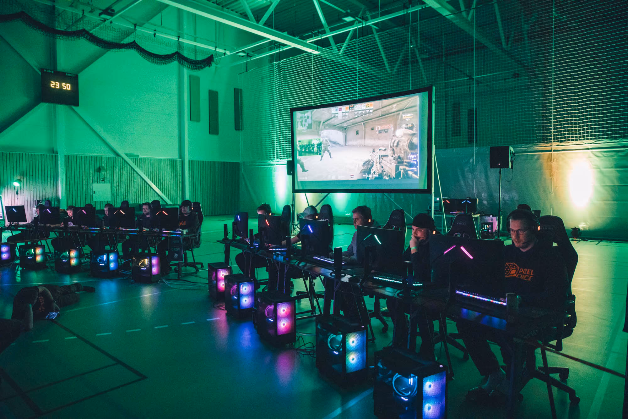 Flere spillere ved datamaskiner konkurrerer i et esport-arrangement med skjermprojektering i en gymsal.