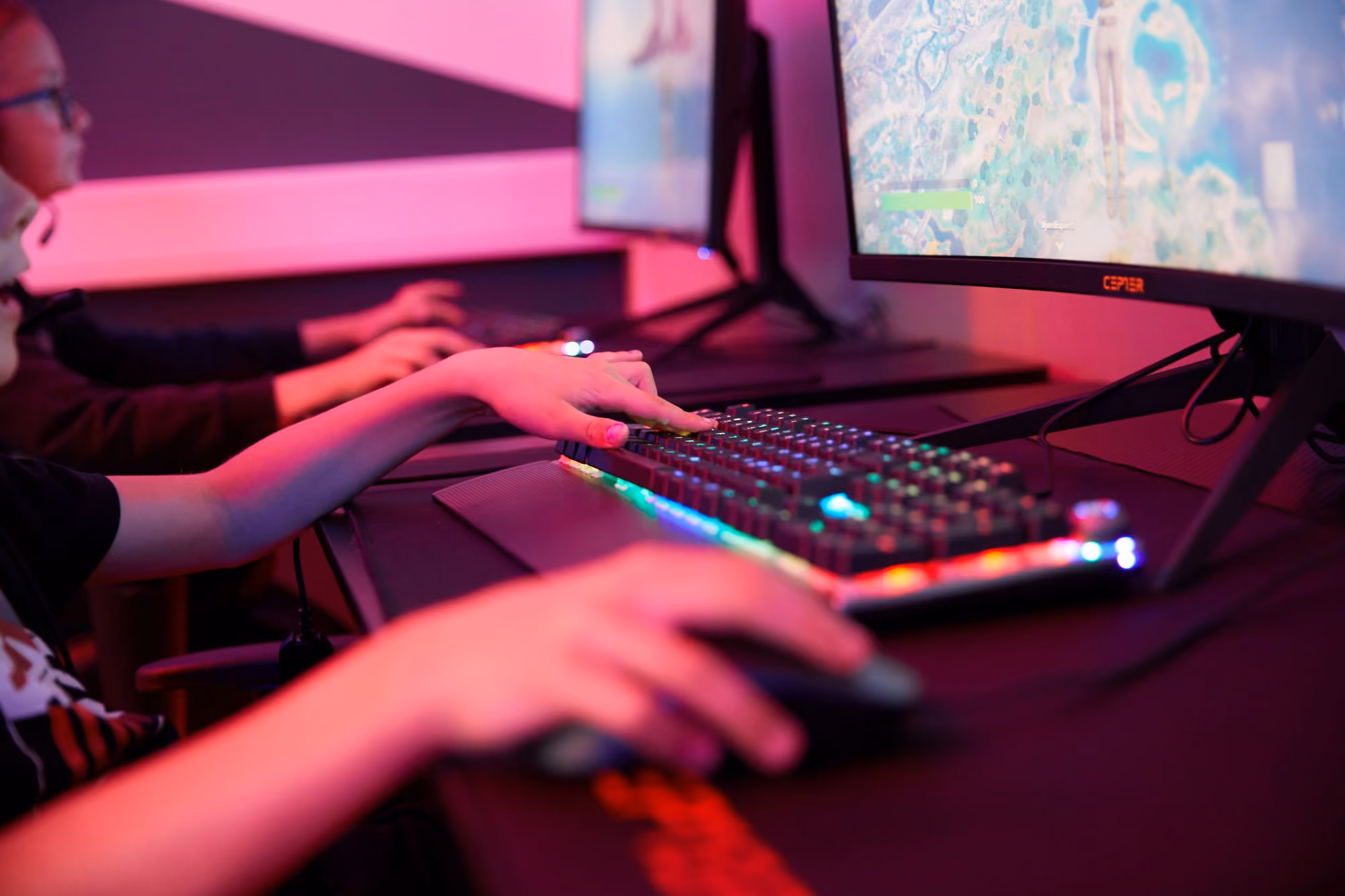 Barn som spiller dataspill med RGB-tastatur og mus foran skjermer med spillgrafikk.