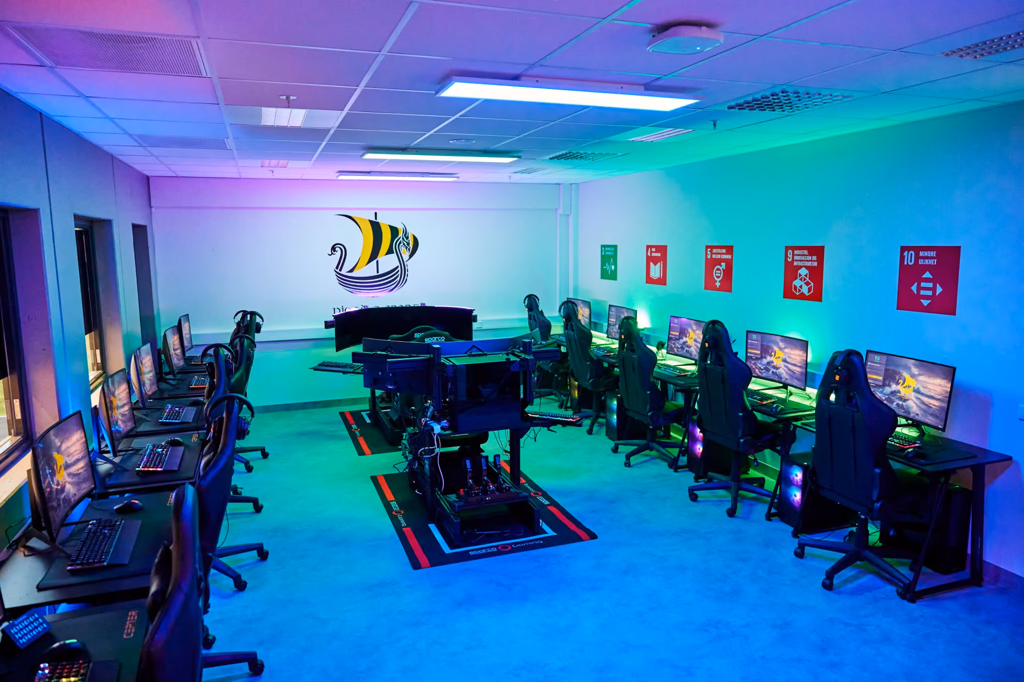 A room with two rows of computers and gaming chairs, with a display setup in the center under blue and green lights.