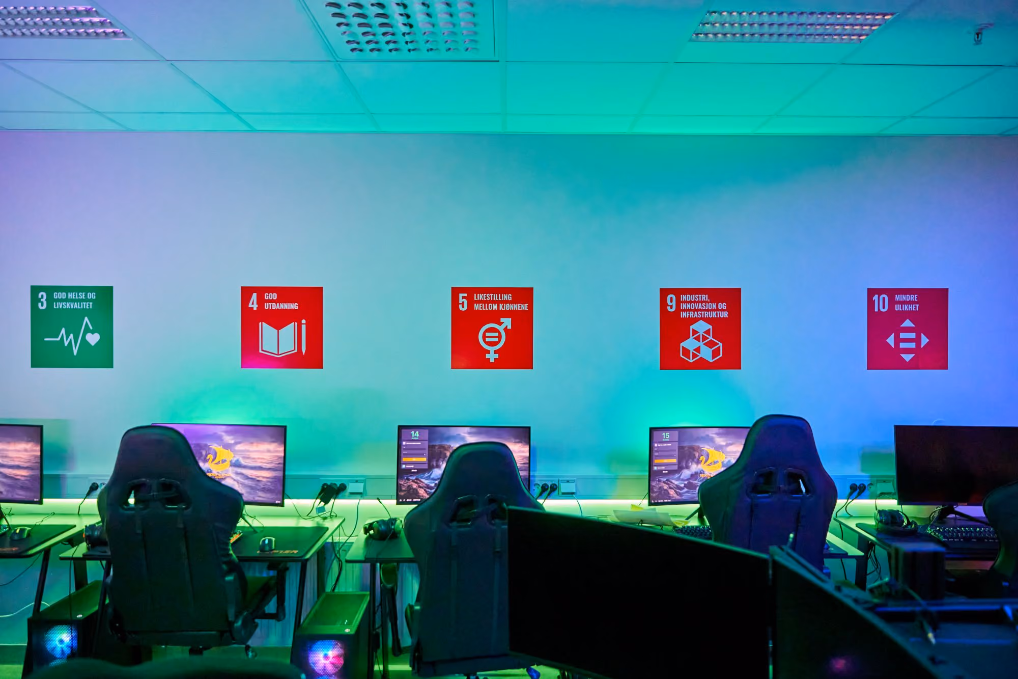 Rom med datamaskiner og gamingstoler under fargerikt lys og bærekraftsmål på veggen.