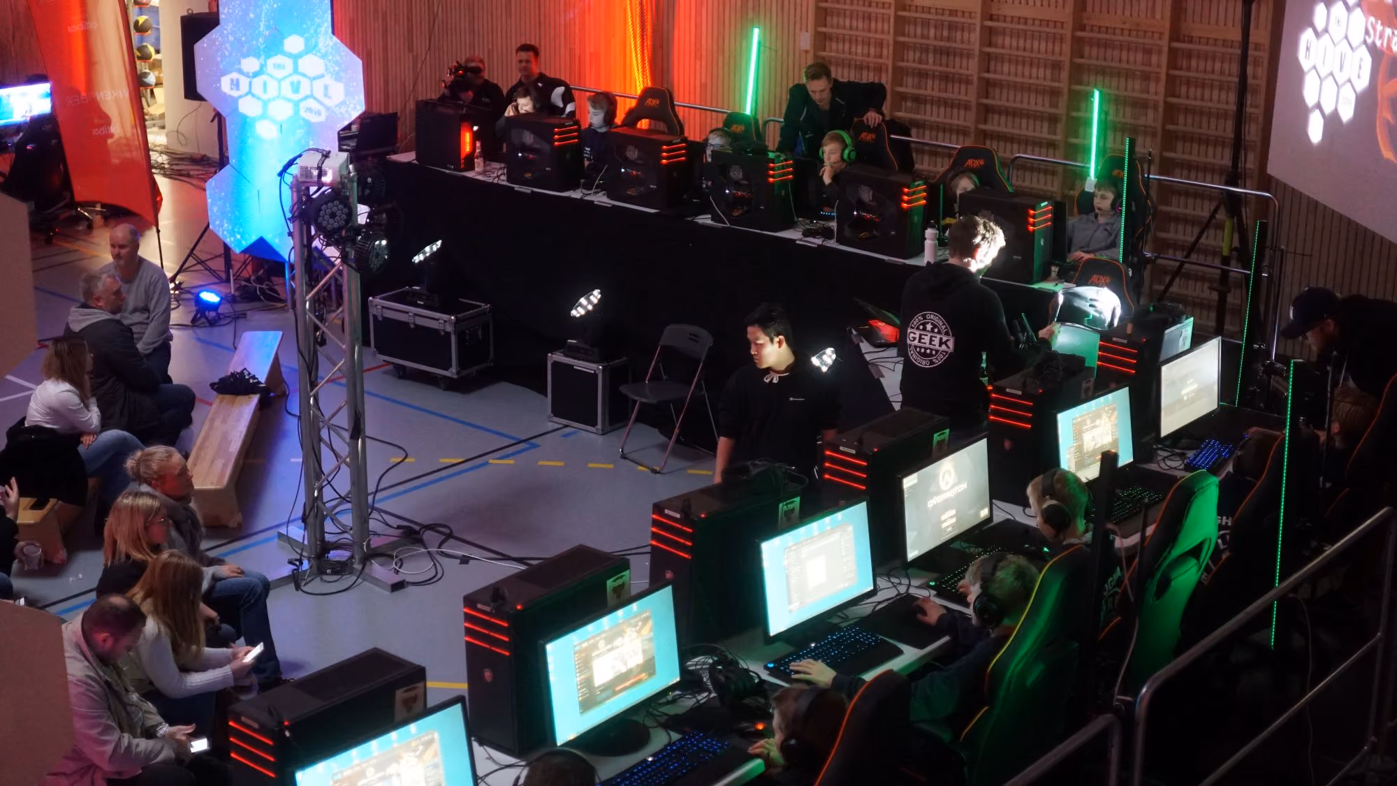 Spillere med hodetelefoner foran skjermer i en esportsarena med publikum på benker.