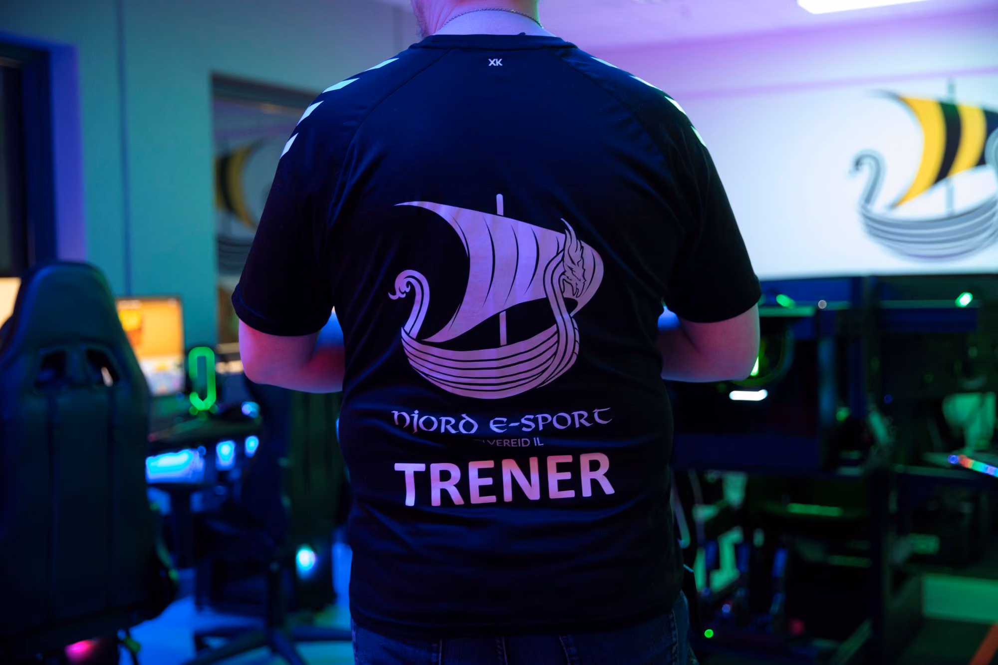 Person med svart skjorte med Nord E-Sport logo og teksten TRENER på ryggen i et gaming-rom.