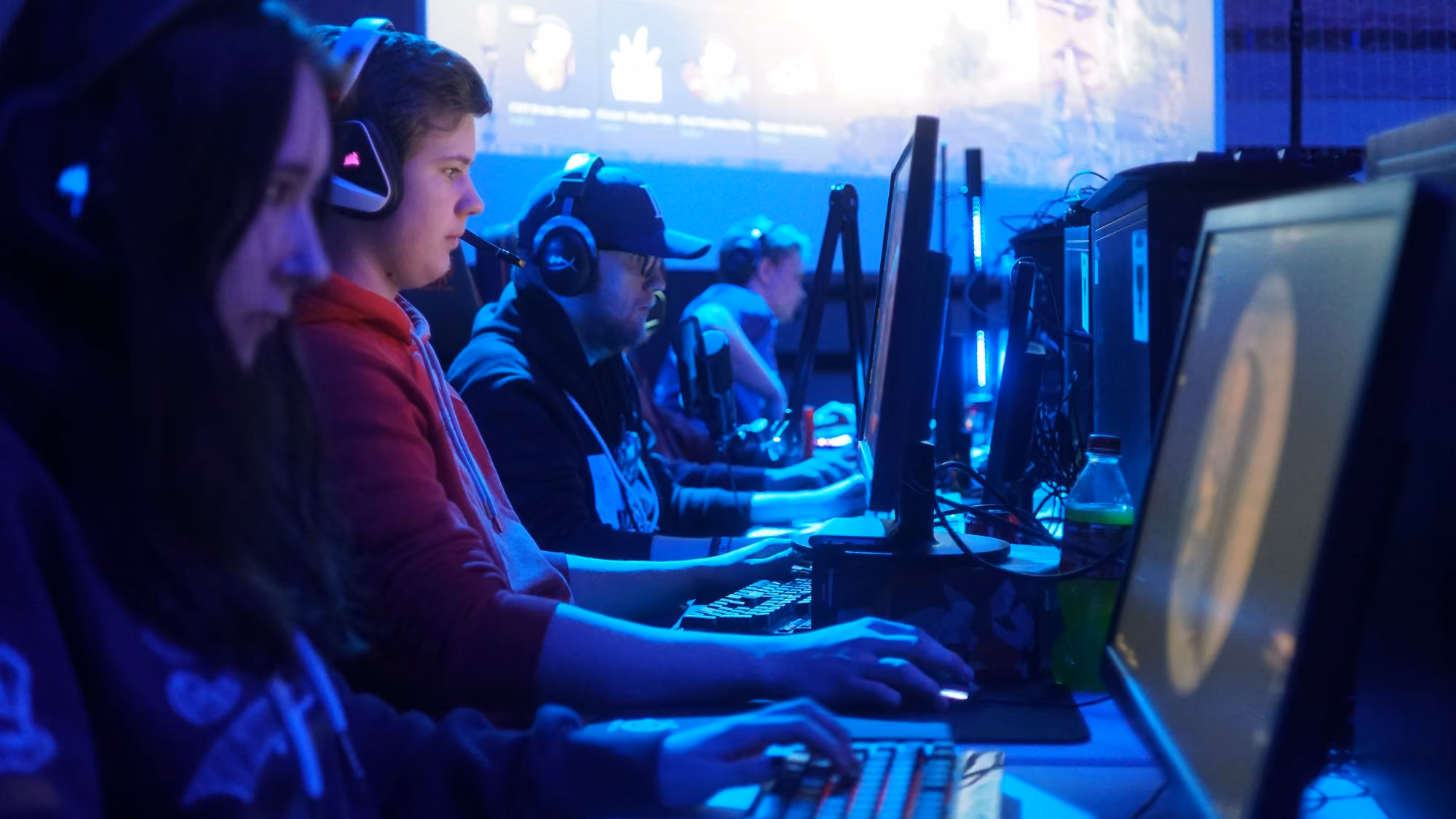 Folk spiller dataspill på PC-er i et rom opplyst av blått lys med skjermer og headset.