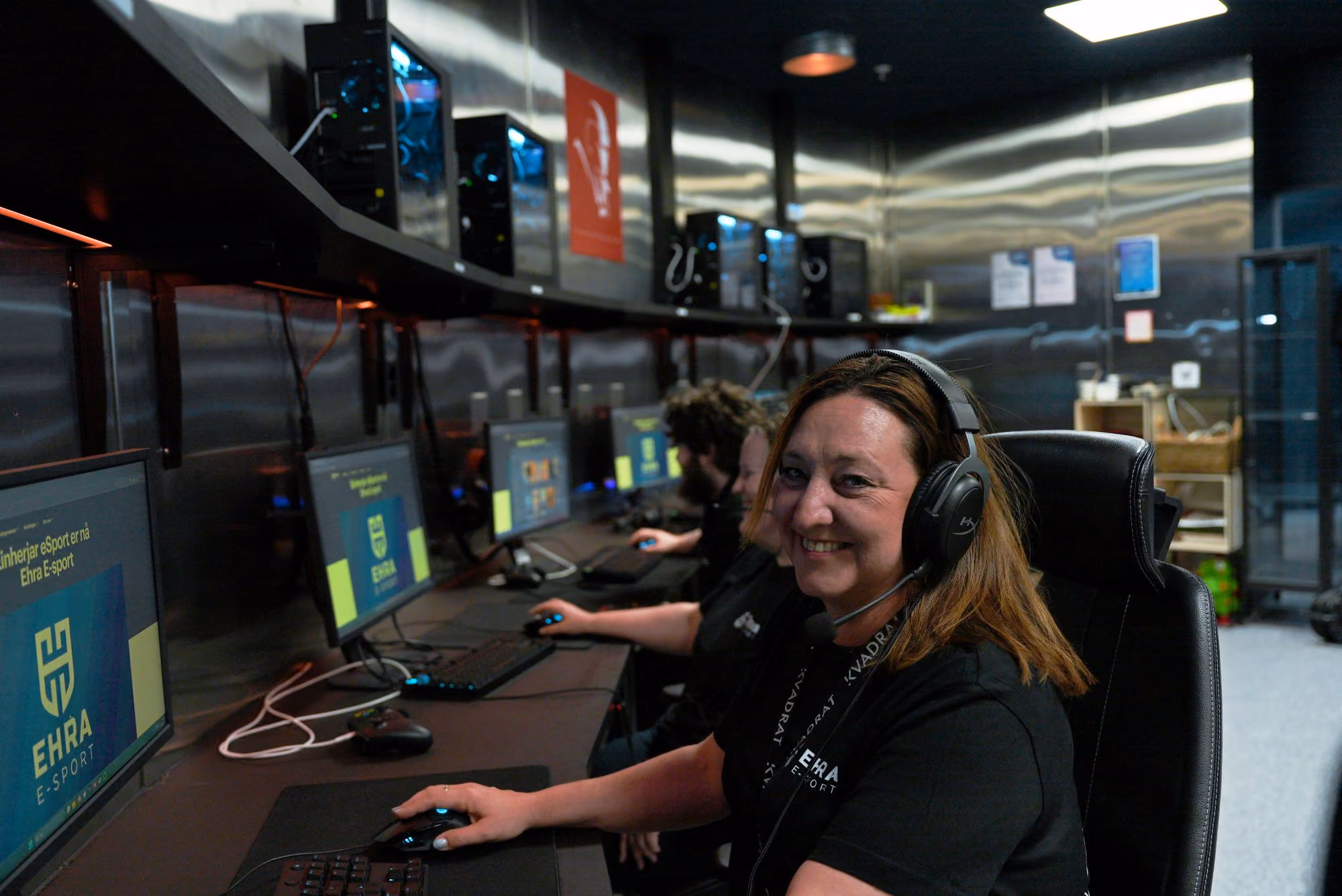 A woman wearing a headset is smiling next to a row of computers in an e-sports room.