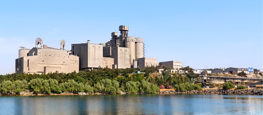 Su kıyısındaki yeşillik ardında yükselen Akçansa çimento fabrikası silo ve kuleleri