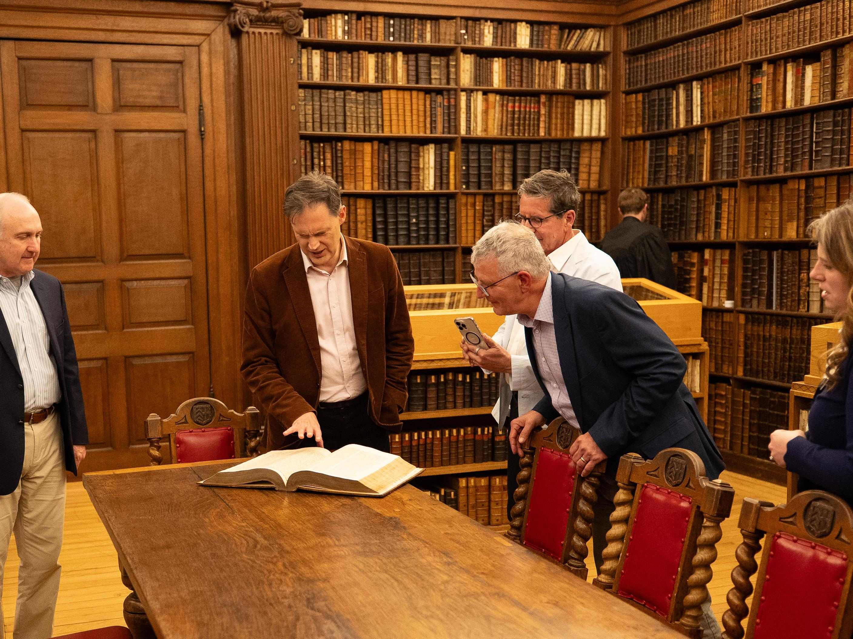 Dr. Peter Williams and visitors examine an open historic Bible in the Fellows’ Library at Clare College, Cambridge, England, surrounded by shelves of rare books.