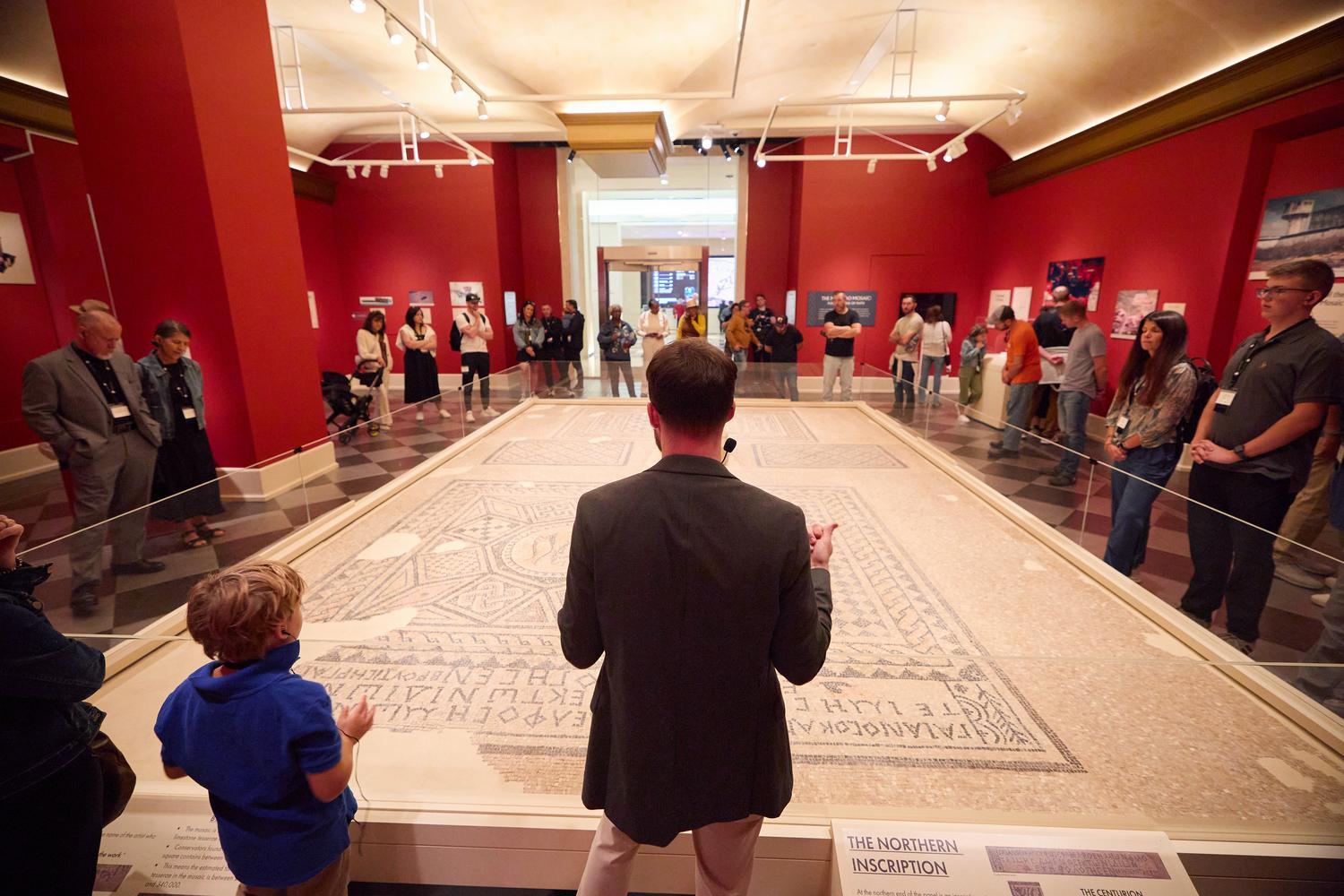 An Inspire guide explains the Megiddo Mosaic as guests gather around one of the earliest known Christian worship mosaics at the Museum of the Bible.