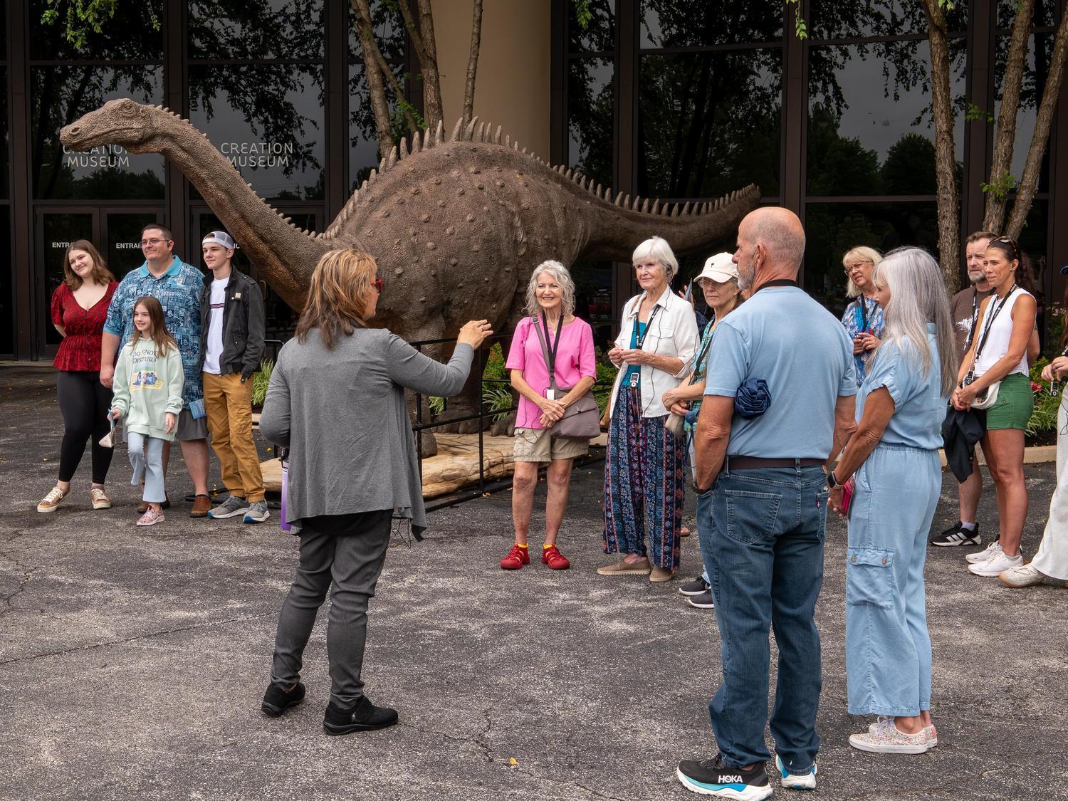 An Inspire guide welcomes guests outside the Creation Museum beside a life-size dinosaur sculpture before beginning a guided tour.