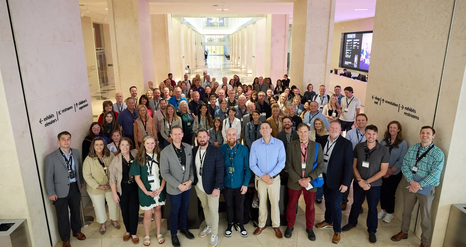Large group of people standing and posing for a photo in a museum lobby with signs on pillars.