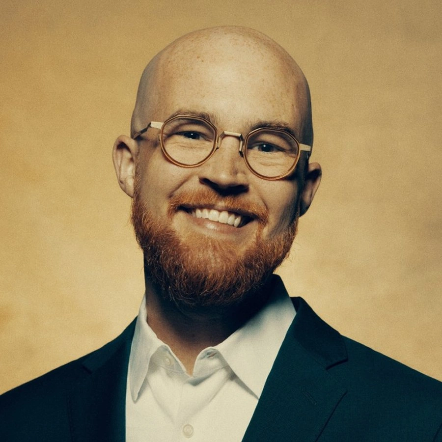 Smiling bald man with red beard and round glasses wearing a white shirt and dark suit jacket.