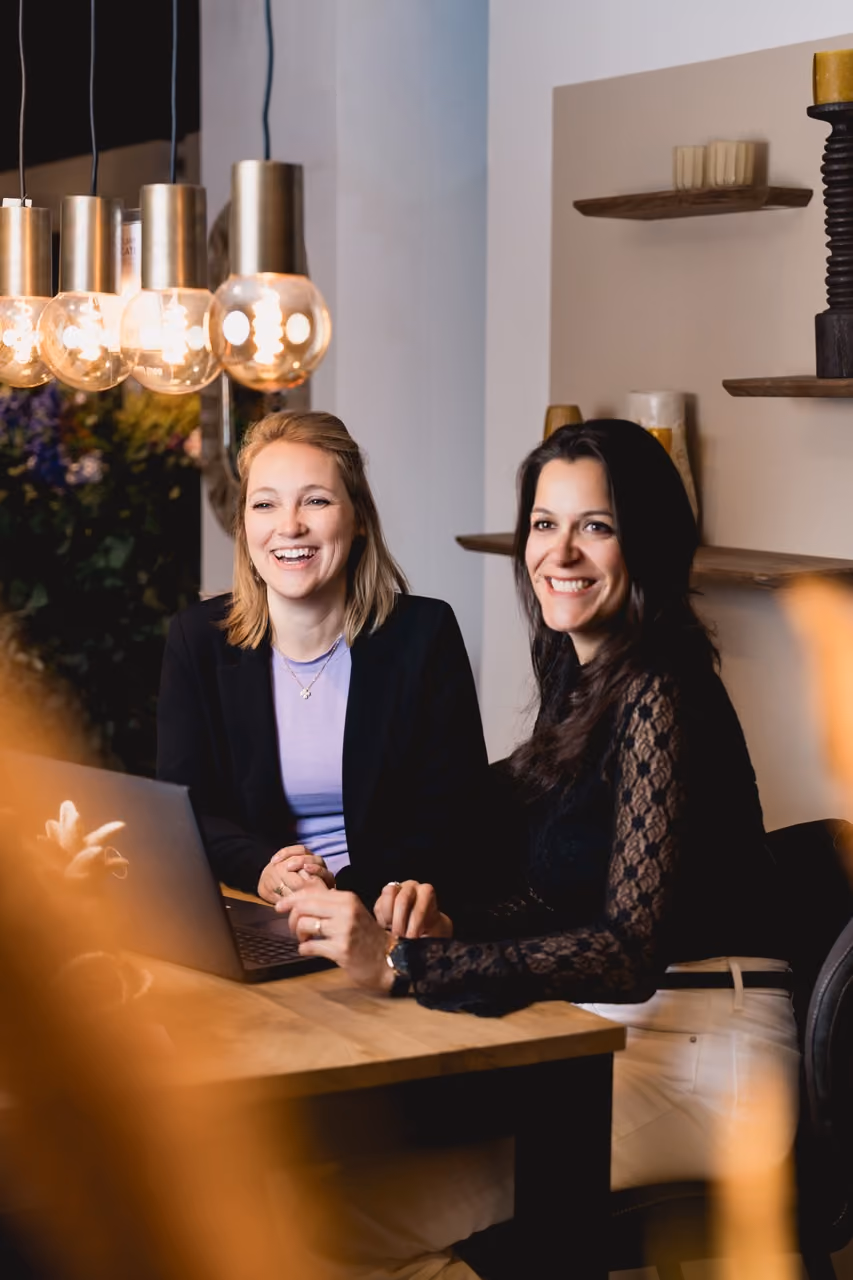 Marlin and Denise sitting at a wooden table with a laptop in a cozy room, smiling and interacting under hanging light bulbs.