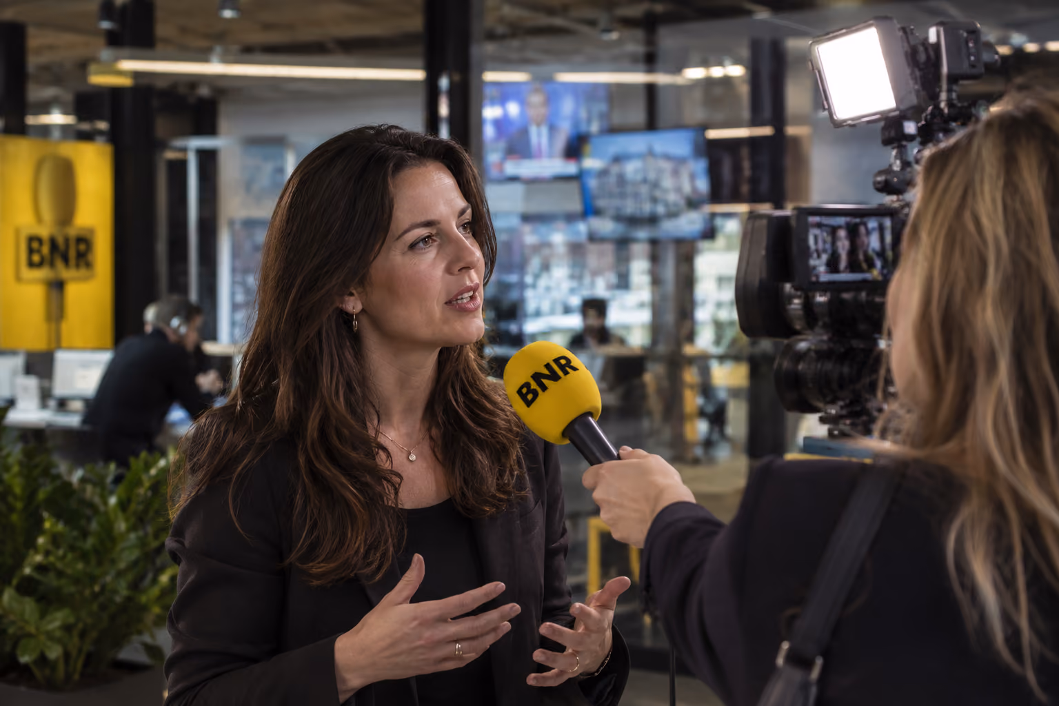 A woman with long brown hair wearing a black blazer is being interviewed indoors, speaking into a yellow microphone labeled BNR while a camerawoman films her with a video camera and bright light.