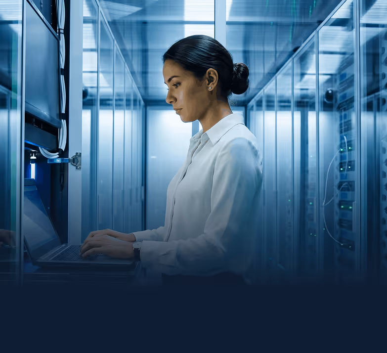 Woman in white shirt working on laptop inside a server room with racks and equipment.