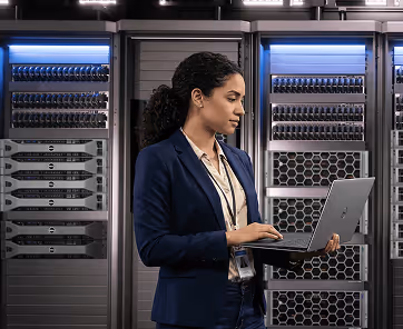 Woman in business attire using a laptop in a server room with racks of hardware behind her.