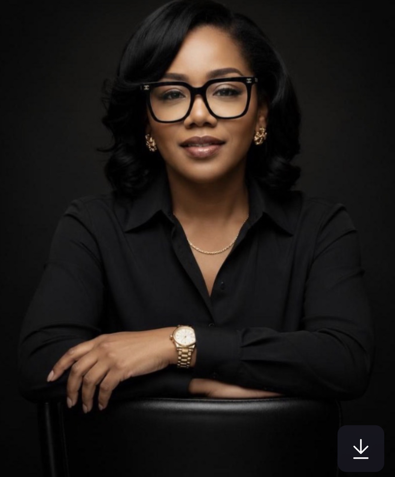 Confident woman with black hair and glasses wearing black blouse, gold watch, and earrings on black background.
