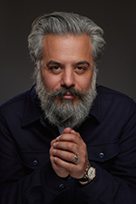 Portrait of a serious man with dark hair and beard wearing a black turtleneck sweater against a gray background.