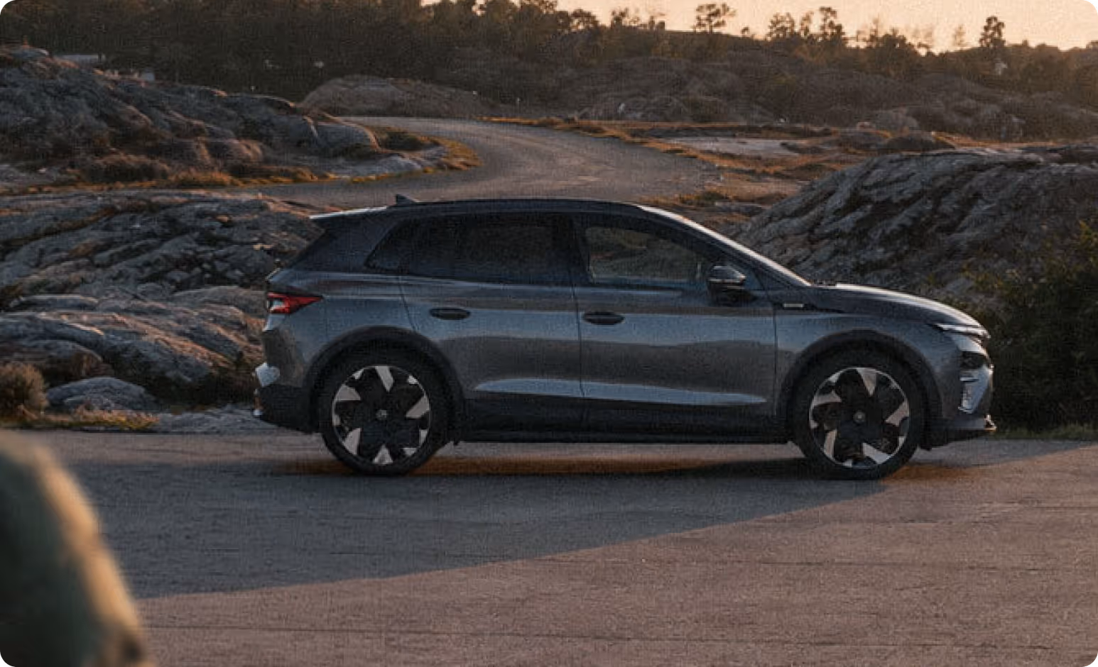Harmaa SUV-auto pysäköitynä kallioiselle alueelle, taustalla mutkainen tie ja puusto.
