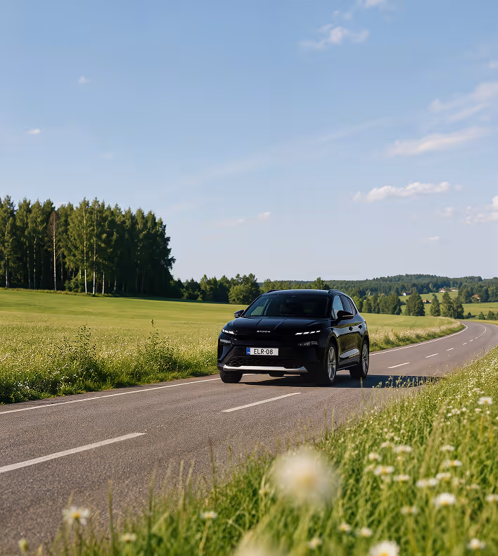 Musta Skoda-auto ajaa asfalttitietä pitkin maalaismaisemassa vihreiden peltojen ja puiden ympäröimänä.