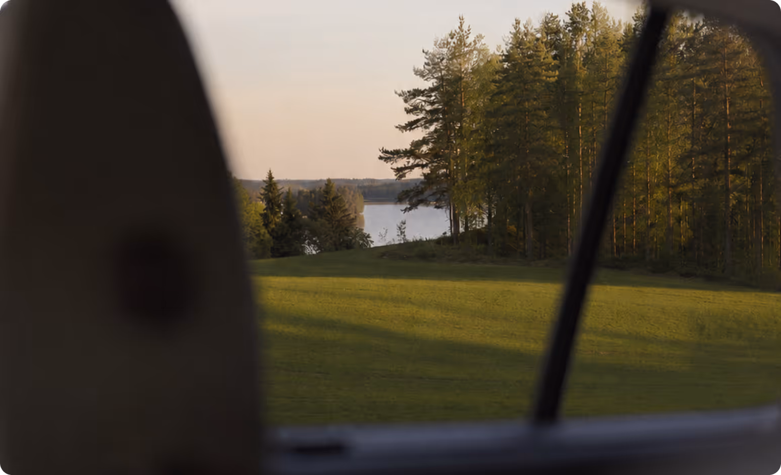 Maisema auton ikkunasta, jossa näkyy nurmikko, mäntyjä ja järvenranta.