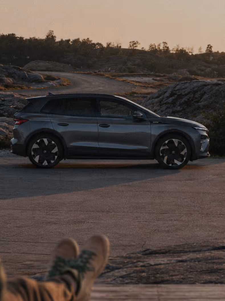 Harmaa SUV seisoo tiellä luonnonkauniissa kallioisessa ja metsäisessä ympäristössä auringonlaskun aikaan.