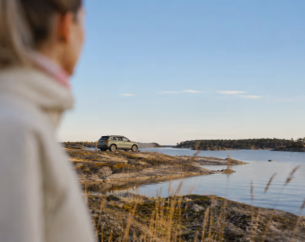 Nainen katsoo harmaata autoa kallioisella rannalla merimaisemassa sinisen taivaan alla.