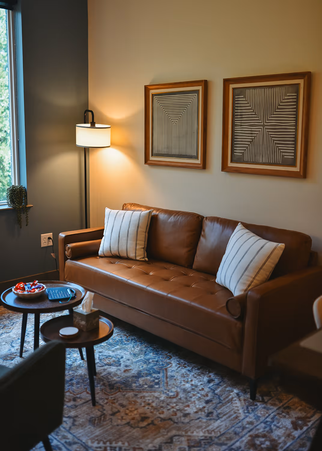 Brown leather sofa with striped pillows next to floor lamp and geometric wall art in cozy living room. New Narratives Counseling